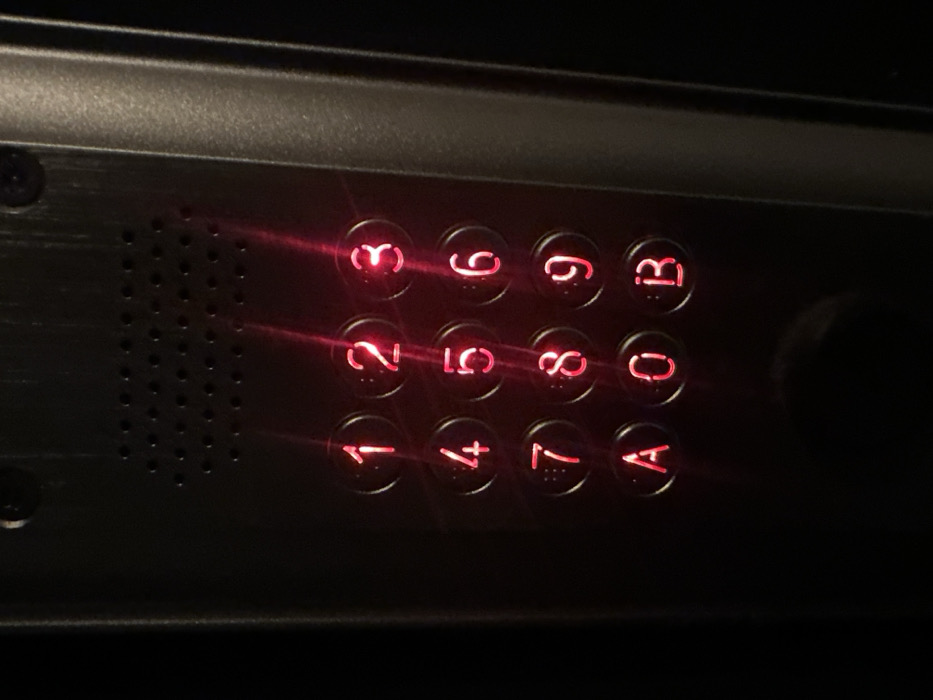 A close-up, low-light photograph displays a metallic access control keypad, likely situated at an entrance point within the city of Paris, France. The keypad is constructed from a dark, possibly brushed aluminum or similar metal, secured by two visible screws at the top corners. Centered above the input buttons is a rectangular grille, perforated with multiple small circular holes, consistent with a speaker or microphone. The keypad comprises twelve circular, illuminated buttons arranged in four rows of three columns. These buttons display red alphanumeric characters: '1', '2', '3' in the top row; '4', '5', '6' in the second; '7', '8', '9' in the third; and 'A', '0', 'B' in the bottom row. Distinct vertical streaks of red light extend across the button surfaces, indicating reflections from the internal LED illumination or ambient light. The surrounding area is obscured by darkness, suggesting a dimly lit indoor environment or nighttime conditions. No individuals or active interactions are visible in the scene.