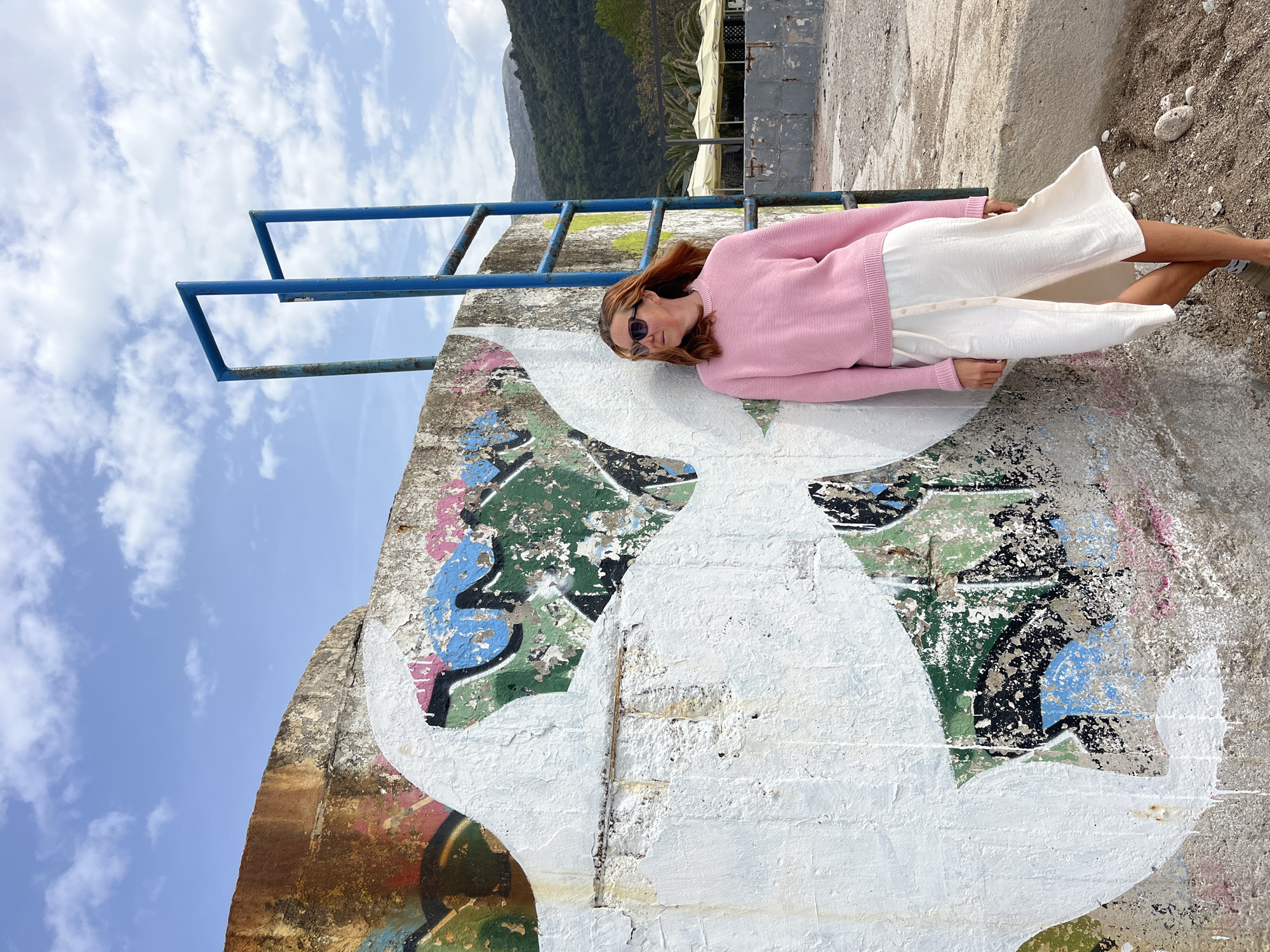 A woman lies on a concrete structure with graffiti in Budva, Montenegro. The woman, wearing a pink sweater, white pants, and sunglasses, reclines against the wall. The concrete structure features layers of graffiti, including white paint covering earlier designs. A blue metal railing is behind the woman. The background includes a mountain, a strip of beach, and palm trees. The sky is blue with scattered clouds. The ground to the right is sandy with rocks.