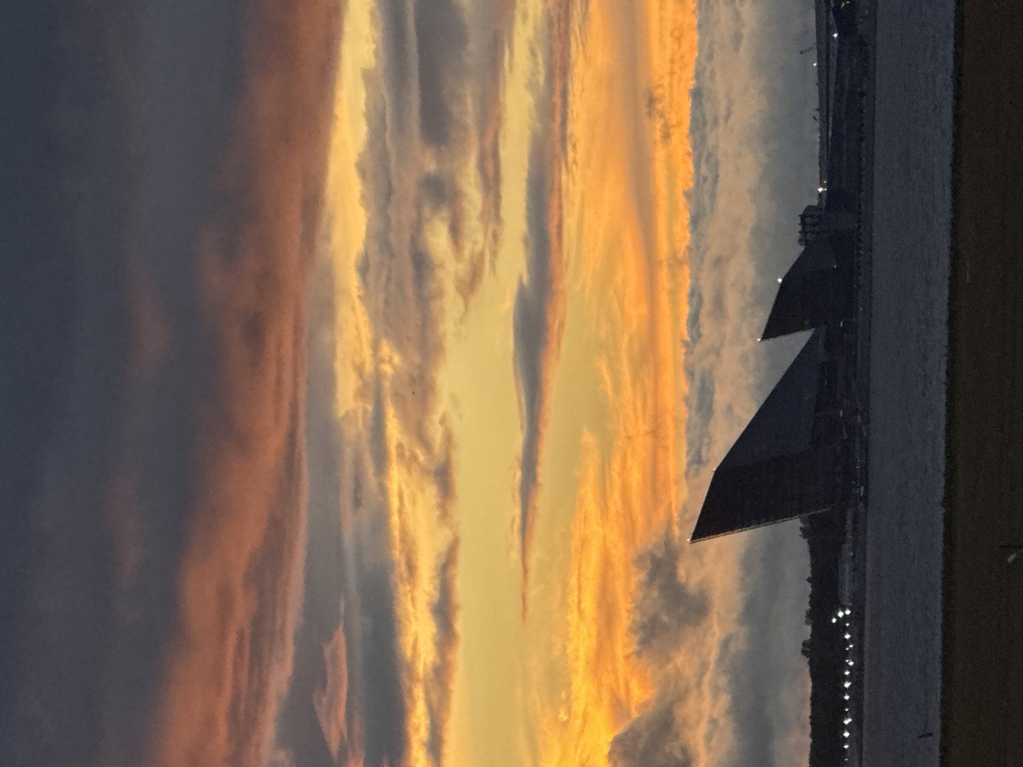 The image depicts a sunset over the Gulf of Finland in Saint Petersburg, Russia. The sky is ablaze with vibrant hues of orange and yellow, transitioning to darker shades of gray and purple at the top. Wispy clouds are scattered across the sky, catching the last rays of the sun. In the foreground, the dark, angular silhouettes of two modern buildings stand out against the water. These are the Lakhta Center and the adjacent Gazprom Arena. The Lakhta Center, the taller of the two, has a distinctive, twisting spire that points towards the sky. The Gazprom Arena, a spherical stadium, is partially visible to the right of the Lakhta Center. The water in the foreground is calm and reflects the colors of the sunset. The overall impression is one of dramatic natural beauty juxtaposed with striking contemporary architecture.
