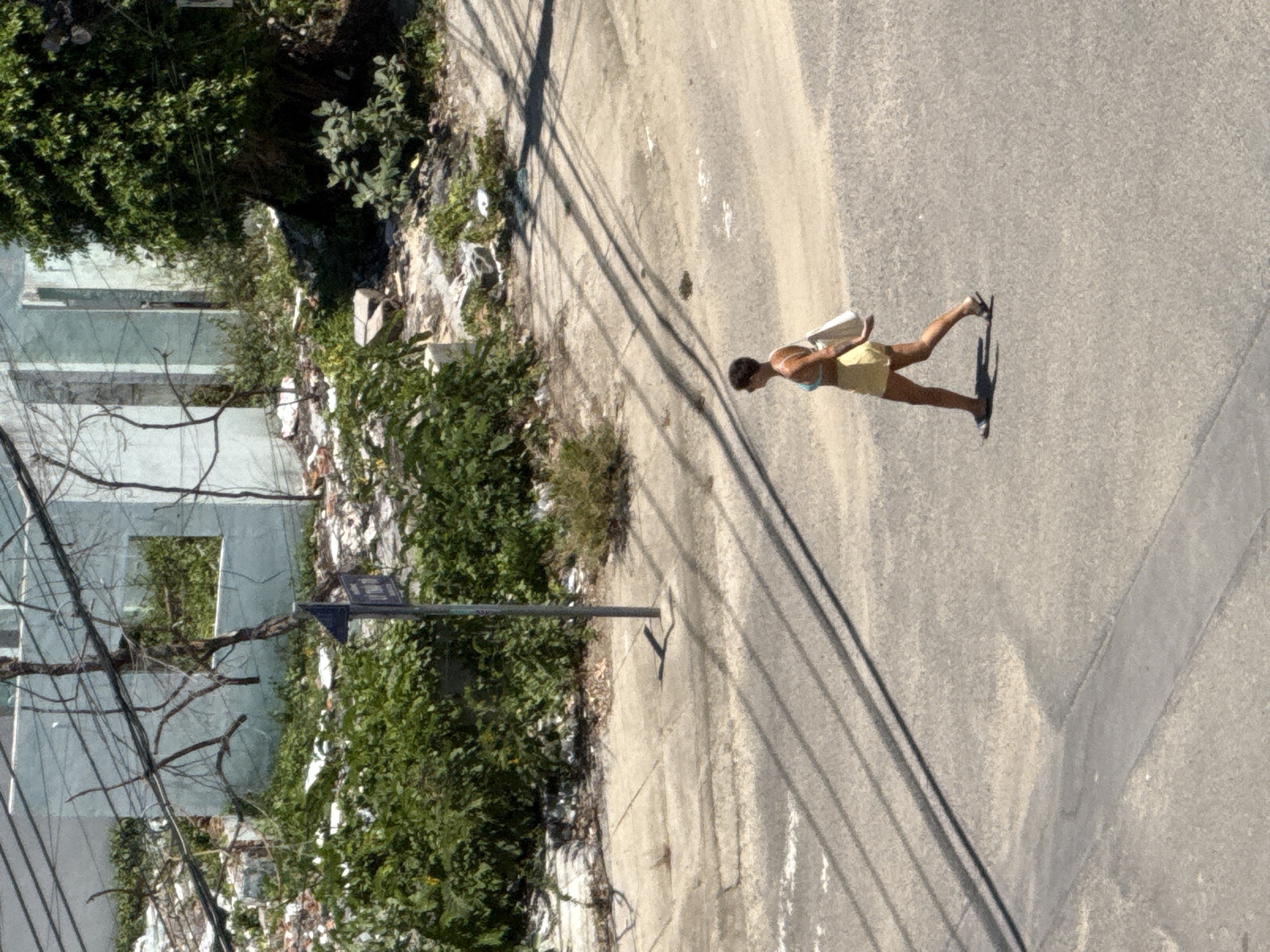 The image shows an outdoor scene in the city of Nha Trang, Vietnam. In the foreground, a woman with short dark hair is walking across an asphalt road from left to right. She is wearing a light blue bikini top and yellow shorts, and carrying a white tote bag over her right shoulder. Her shadow is cast on the road behind her. The road has some painted white lines and a shadowed pattern from overhead wires. In the background, a dilapidated building with light blue walls and broken windows is visible, surrounded by overgrown green vegetation and debris. A utility pole with a triangular sign is located on the sidewalk in front of the building. The overall impression is of a somewhat neglected urban environment.