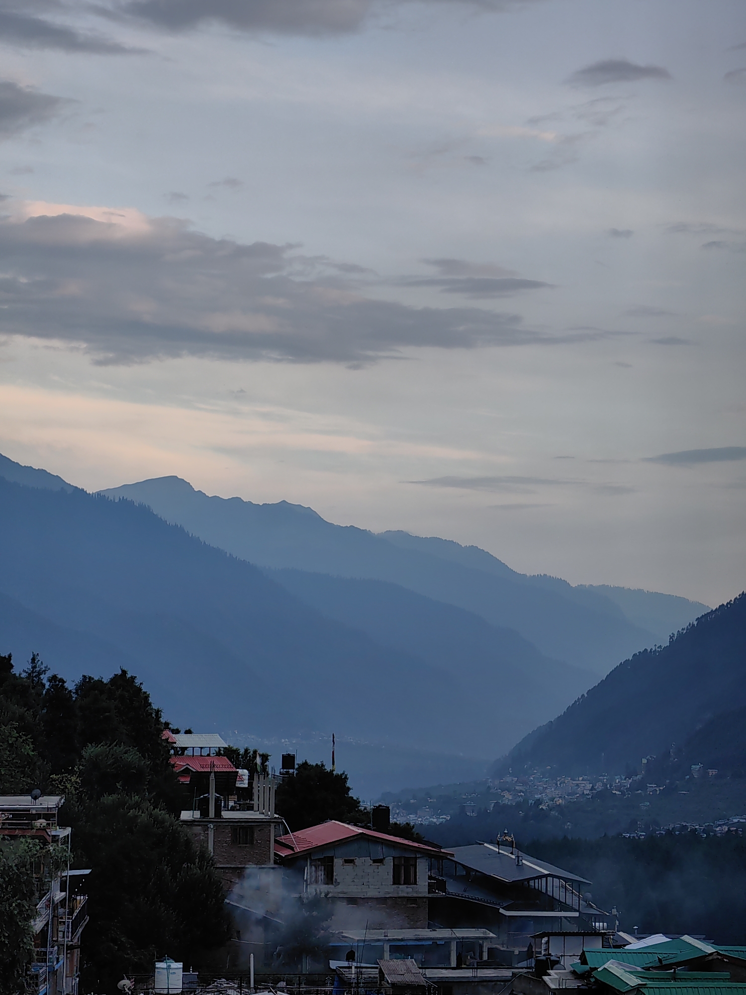 An aerial, medium shot captures the misty twilight over a village nestled in a valley, with layered blue mountains receding into the distance. The sky is a blend of pale blue and gray clouds, with hints of pink and orange near the horizon. In the foreground, several buildings with red and green roofs are visible, interspersed with lush green trees. Smoke drifts from some of the structures, adding to the atmospheric haze. The town extends up the mountainside, with lights visible in the distance. The overall impression is one of tranquility and natural beauty, with a hint of human habitation blending seamlessly into the landscape. The image is an exterior shot of the village of Vashisht, India.