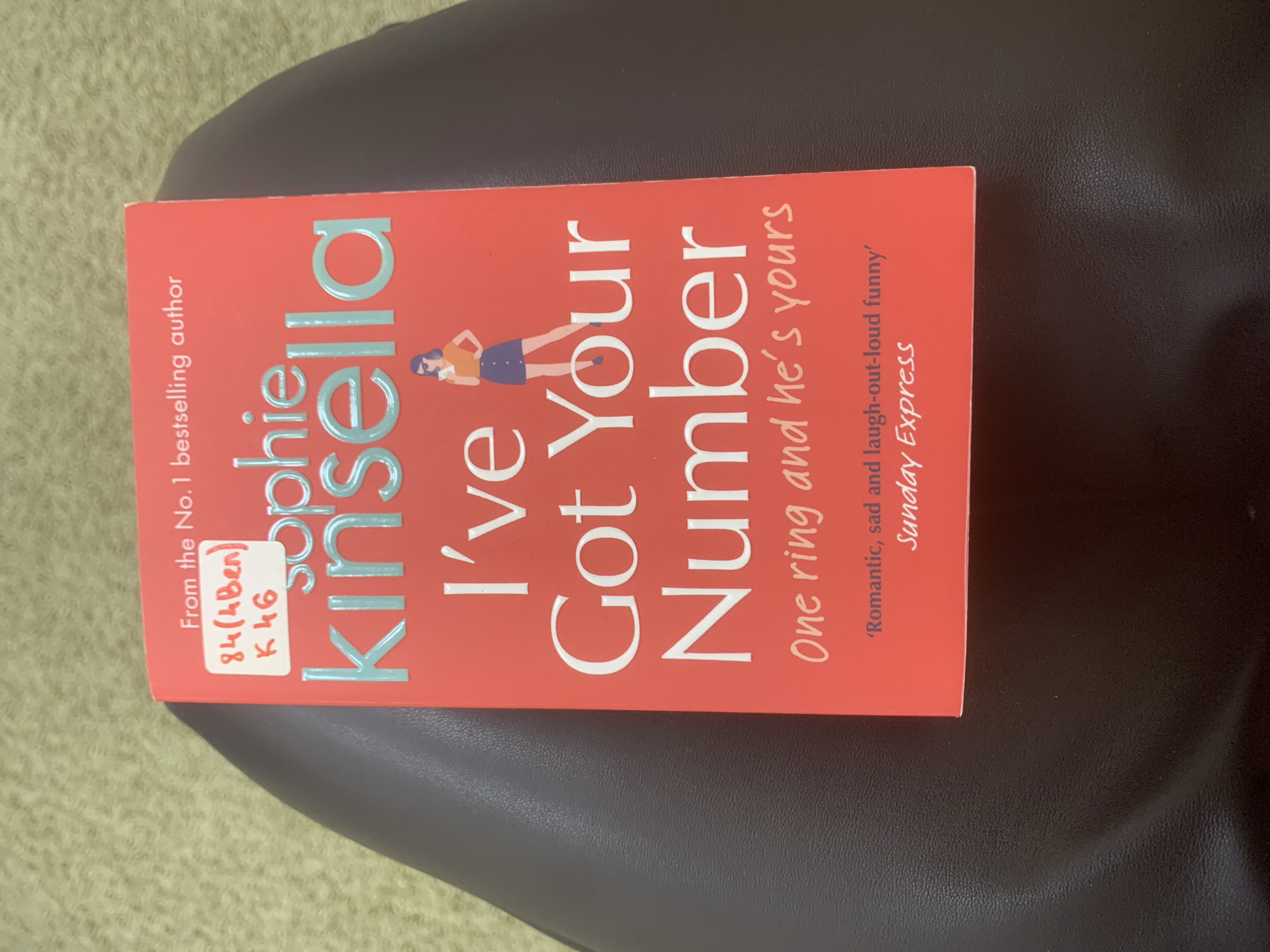 The image shows a paperback copy of the book "I've Got Your Number" by Sophie Kinsella. The book has a red cover with the title prominently displayed in white text. An image of a woman is shown between the words "I've" and "Got." The cover also includes the tagline "One ring and he's yours" and a quote from the Sunday Express. There's a library sticker in the upper left corner with the number "84(4Ben) K46". The book is lying on a dark brown surface that appears to be a chair. The background is blurry with hints of light green. There's no indication of the image being taken in Izhevsk, Russia.