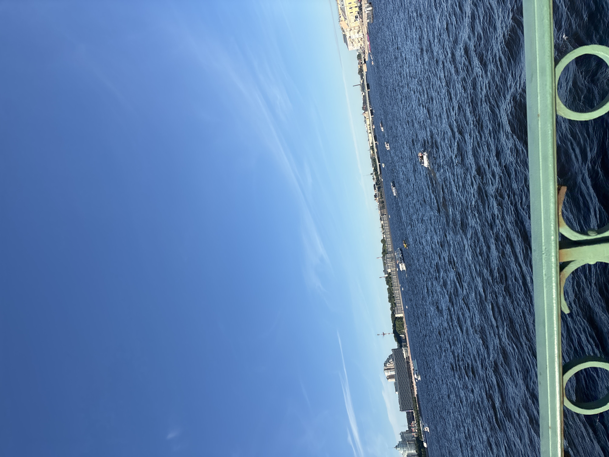 The image captures a wide view of the Neva River in Saint Petersburg, Russia, on a clear, sunny day. The deep blue water is choppy with visible waves, reflecting the bright blue sky dotted with thin, wispy clouds. In the foreground, the ornate green railing of a bridge or embankment is visible, with decorative circular patterns.
Across the river, the St. Petersburg skyline is lined with buildings of various architectural styles, including modern apartment blocks and older, more classical structures. Several boats of different sizes, including what appear to be tour boats and smaller personal vessels, are scattered across the river. One small white boat in the mid-right of the frame has at least two people aboard. The city's urban landscape extends into the distance, with bridges visible in the background. The overall scene depicts a lively urban waterway on a pleasant day.