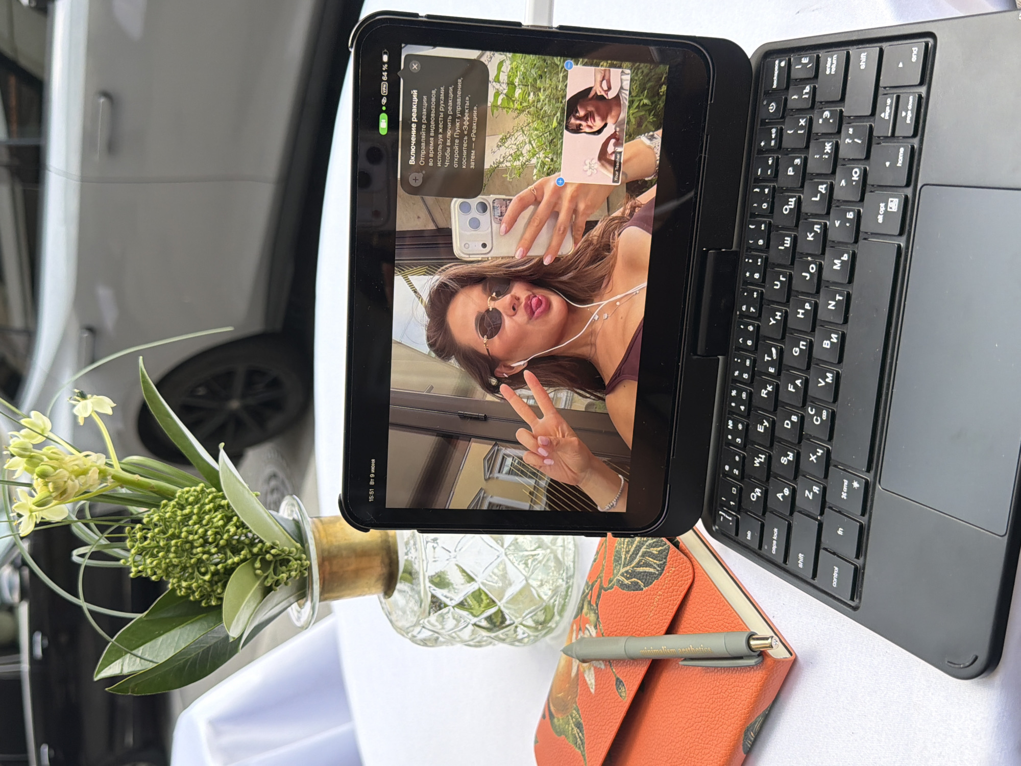 The image depicts an outdoor setting, likely a cafe or restaurant, with a white tablecloth on a table. In the foreground, a tablet with a keyboard attached is open, displaying a video call. The main participant in the video call is a woman taking a selfie with her phone, making a peace sign and sticking her tongue out, while wearing sunglasses and headphones. A smaller video feed shows another woman with her hand near her face. The tablet screen also shows system information at the top, including the time "15:51" and the date "9 ИЮНЯ" (June 9th), along with battery status and VPN connection.
To the left of the tablet, there is an open notebook with an orange cover featuring a floral design, and a gray pen resting on it. Behind the tablet, a glass vase with a floral arrangement is visible, consisting of green leaves and light-colored flowers. In the background, a blurred image of a silver car and some greenery can be seen. The presence of the time and date on the tablet, coupled with the style of the car and the fashion of the woman, suggests a contemporary urban environment. While the image does not explicitly state the location as Saint Petersburg, Russia, the Cyrillic text on the tablet screen indicates a Russian-speaking context. The overall scene conveys a casual and social interaction taking place outdoors.