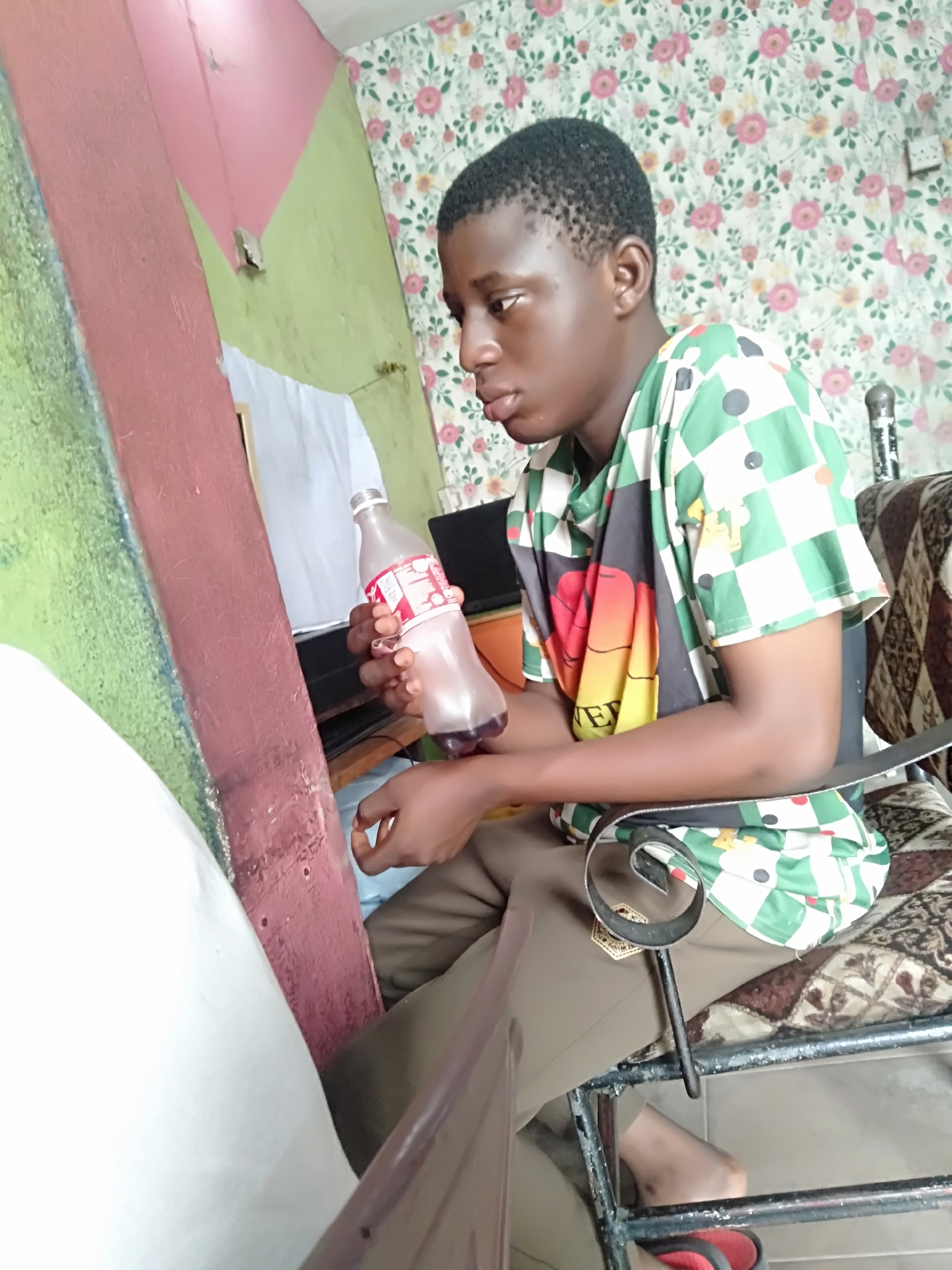A young man is sitting in a metal chair, indoors. He is holding a partially empty plastic bottle with a red label. He is wearing a green and white checkered shirt with a colorful design in the center. He has short, dark hair and brown skin. His pants are khaki colored. The background includes a floral wallpaper, a computer monitor, and a wall painted green and pink. The location is in Shangisha, Nigeria.