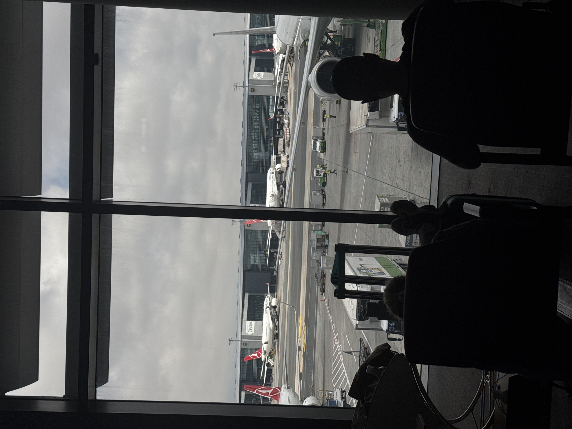 The image depicts an airport scene viewed from inside a terminal, likely a lounge area, looking out through large windows. In the foreground, two figures are silhouetted in chairs, facing away from the camera and towards the exterior view. The left figure is seated, with their legs visible, and a piece of luggage to their side. The right figure is also seated, facing the window.
The exterior scene reveals an airport tarmac with multiple airplanes. One prominent airplane, white with red tail fins bearing a Turkish Airlines logo, is parked on the tarmac. Other aircraft are visible in the distance. The airport terminal building, a modern structure with large glass windows, stretches across the background. Various airport service vehicles and equipment, such as baggage carts and service trucks, are present on the tarmac, with some airport personnel visible wearing high-visibility vests. The sky is overcast, suggesting a cloudy day.
This scene is consistent with the Istanbul Airport (IST) located in Arnavutköy, Turkey, known for its modern facilities and extensive operations, including numerous Turkish Airlines flights.