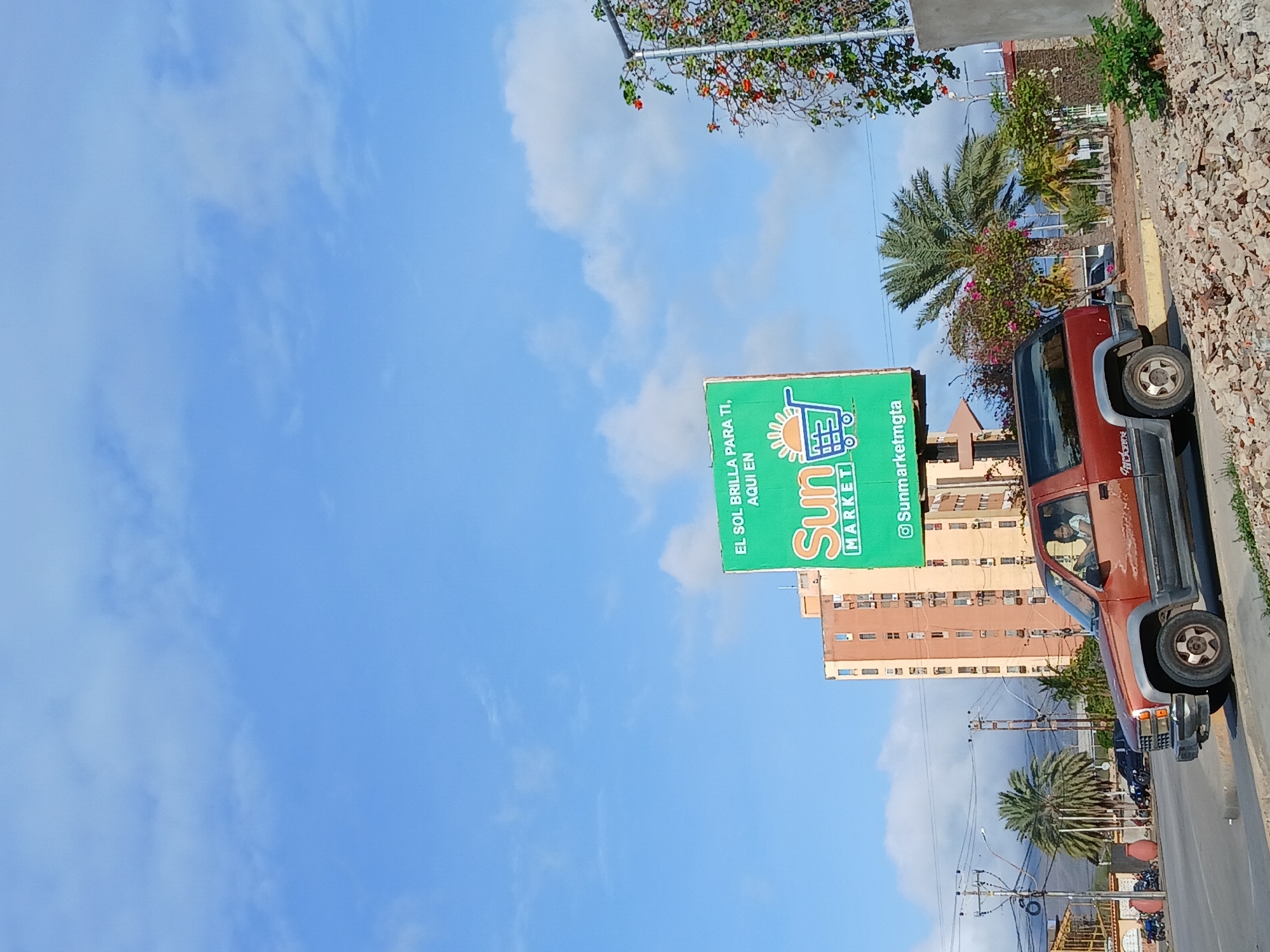 The image depicts a bright, sunny day in Juan Griego, Venezuela. The sky is a clear blue with scattered white clouds. In the foreground, a rusty brown Chevrolet SUV is parked on the side of a road, with its driver, a young female with light-colored hair, visible behind the steering wheel. The SUV is facing to the left. Behind the SUV, a tall, multi-story building with many windows is visible, colored in shades of orange and beige. To the right of the building, a large green billboard is prominently displayed, advertising "Sun Market" with the slogan "EL SOL BRILLA PARA TI, AQUI EN". A shopping cart icon is depicted on the billboard. Further to the right, a street lamp is adorned with red flowers. The environment appears to be urban, with palm trees and other vegetation visible. The ground is covered with gravel and debris in the immediate foreground. Other vehicles and buildings are visible in the background, suggesting a bustling city scene.