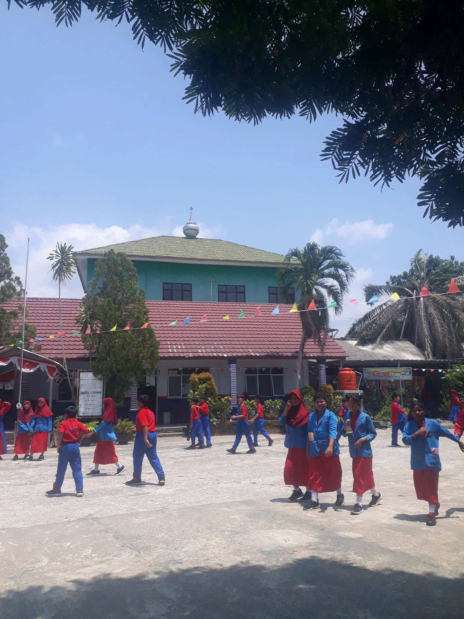 In Pangkalpinang, Indonesia, on a clear, sunny day, a group of uniformed students, some wearing red shirts and blue pants and others blue jackets and red skirts, are gathered on a paved outdoor area. The students appear to be participating in a school activity or event. A two-story building with a green roof and a dome-like structure on top is visible in the background, possibly a mosque or a school building. Red-tiled roofs and colorful triangular flags adorn the buildings. Several trees, including palm trees and a tall evergreen, are scattered around the area. The scene suggests a typical school day in Pangkalpinang, with students engaged in outdoor activities under the bright Indonesian sun.