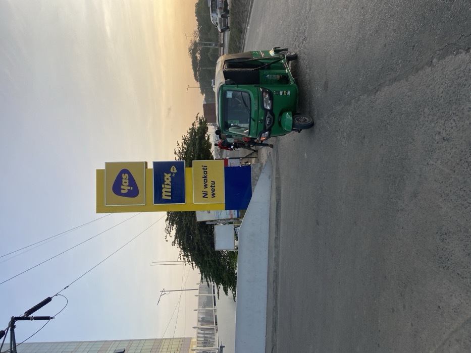 The image shows a green three-wheeled auto-rickshaw parked on the side of a road in Dar es Salaam, Tanzania, at the coordinates -6.779, 39.218. Two people are standing nearby. The auto-rickshaw is facing towards the camera. In the background, there are power lines, street signs, and a building. One sign advertises the words "Mix" and "Yas" in white on a yellow background. Another sign is in Swahili and reads "Ni wakati wetu." There are trees on both sides of the street. The sky is a light blue with a few white clouds and the sun is setting in the distance. The road is paved with asphalt and appears to be in good condition. The environment is a typical urban scene.