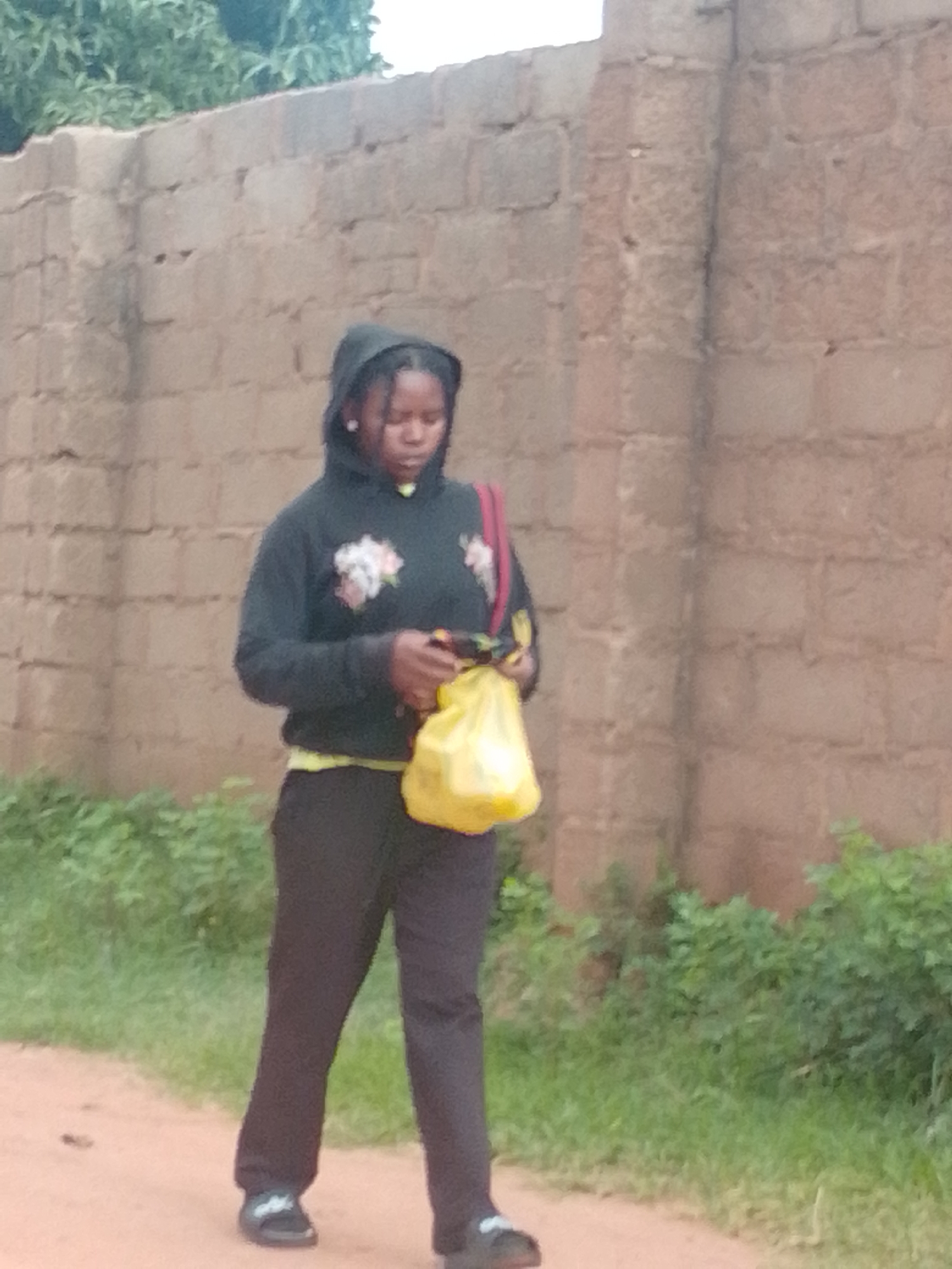 A young Black person wearing a dark hoodie with floral designs and black pants walks along a dirt path next to a brick wall and overgrown green vegetation. They are carrying a bright yellow plastic bag in their right hand. The person appears to be walking away from the camera, with their head down. The background consists of a light-colored brick wall and lush green foliage, suggesting an outdoor setting, possibly in a less urbanized area within Gombe, Nigeria. The lighting is neutral, indicating it is daytime.