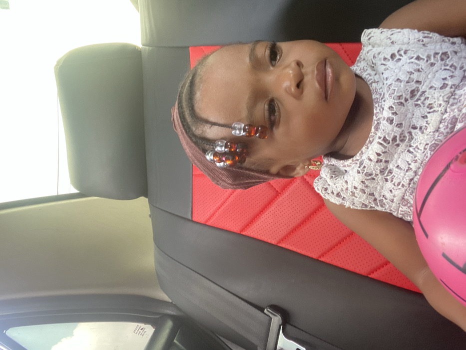 A young child is seated in the rear passenger seat of a vehicle in Adankolo, Nigeria. The child, with dark skin and dark eyes, maintains a neutral expression, gazing slightly to the right of the frame. They wear a dark reddish-brown headwrap, beneath which braided hair with clear and amber beads is visible on the forehead. Small gold-colored hoop earrings are present. The child is dressed in a light-colored, sleeveless top with a distinctive lace or crocheted pattern. A bright pink ball rests in the child's lap. The vehicle's seat features black upholstery with a red, perforated, quilted central insert. A black headrest is positioned behind the child, and a black seatbelt is visible on the left side of the frame. Bright daylight illuminates the scene, visible through the vehicle's window.