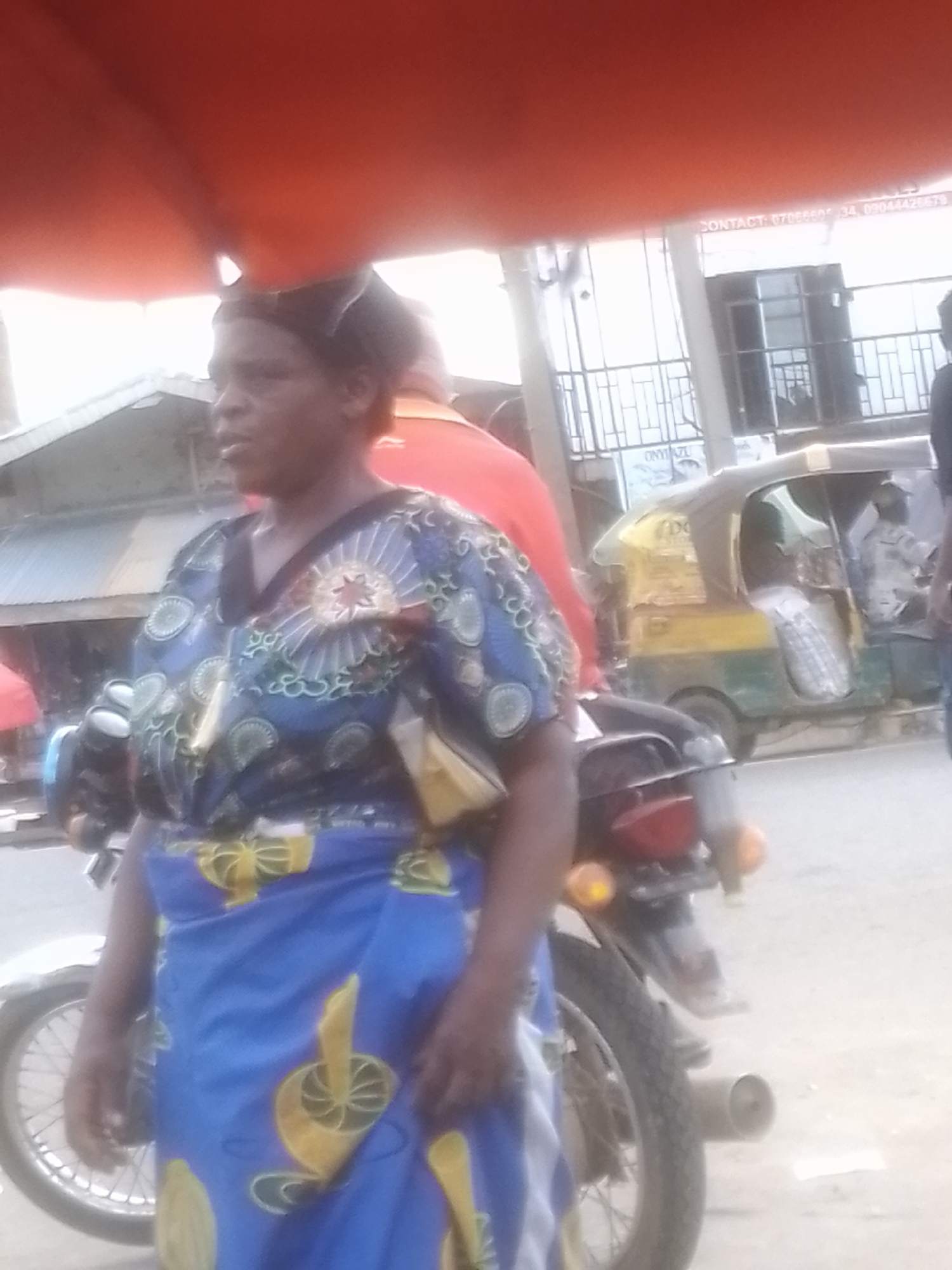In Gwong, Nigeria, a woman stands in front of a motorcycle, her body turned slightly to the left. She is wearing a vibrant blue and yellow patterned dress, with the fabric of her skirt featuring large yellow circular motifs. A smaller yellow bag is tucked into her waist. The background shows a street scene, with a tuk-tuk vehicle visible to the right of the woman. The tuk-tuk is primarily yellow and green, with its doors open. In the distance, there appears to be a building with a metal gate or fence. The overall lighting suggests it is daytime, likely overcast or hazy. The woman's expression is neutral. Visible text on a sign in the background reads "CONTACT: 07066605 34, 09044426879". Another partial text on a sign beneath the building reads "ONYIAZU".