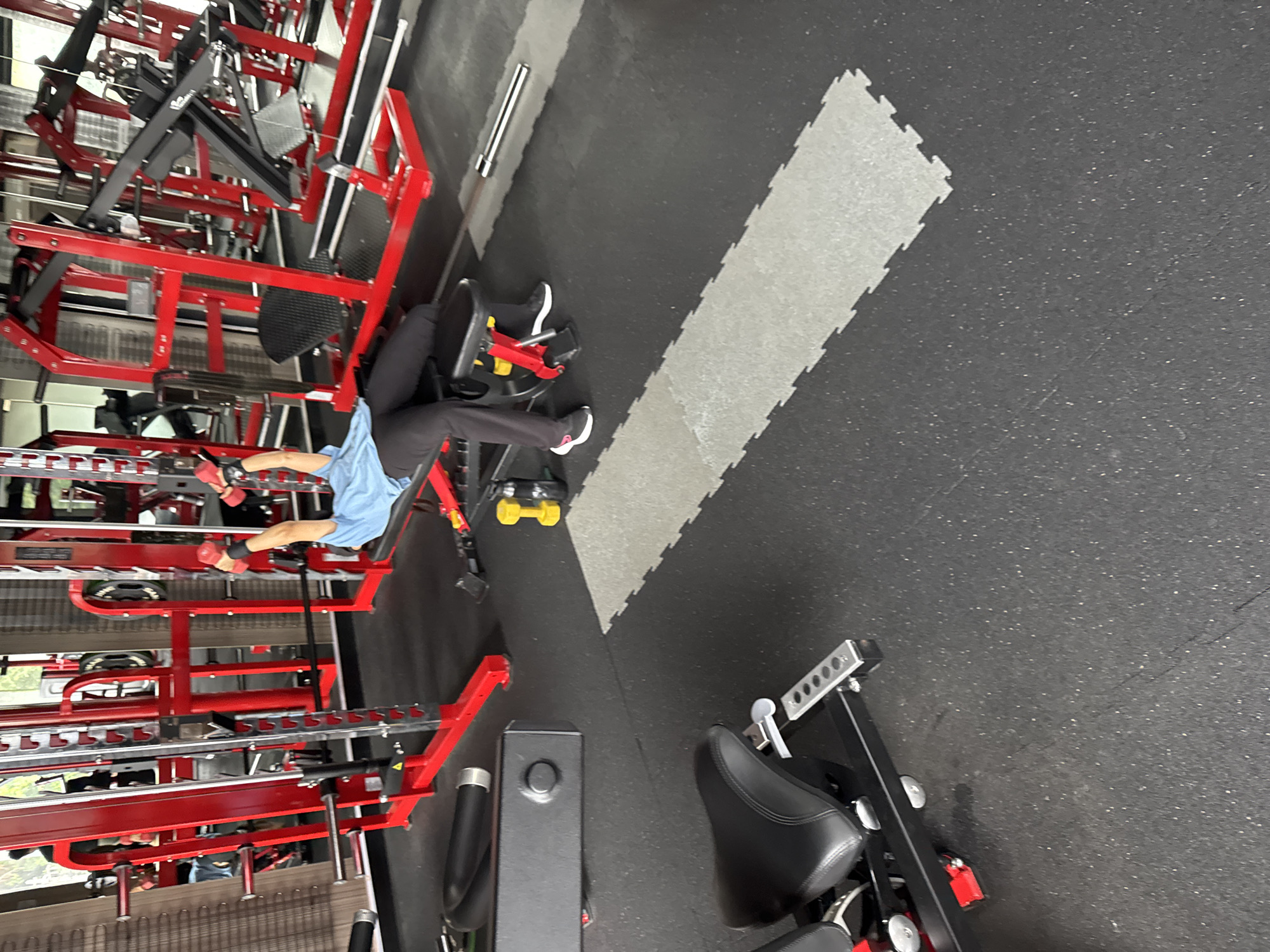 A person is performing a dumbbell exercise on a bench press in a gym. The person is wearing a light blue t-shirt and black pants. They are holding a dumbbell in each hand and their arms are extended above their chest. The bench press is red and is part of a larger gym setup with other red and black weight machines. The gym floor is black with some grey interlocking mats. The background shows more gym equipment and reflections in mirrors. The image appears to be taken in a modern, well-equipped gym. No specific location in Kolkata, India can be determined from the image.