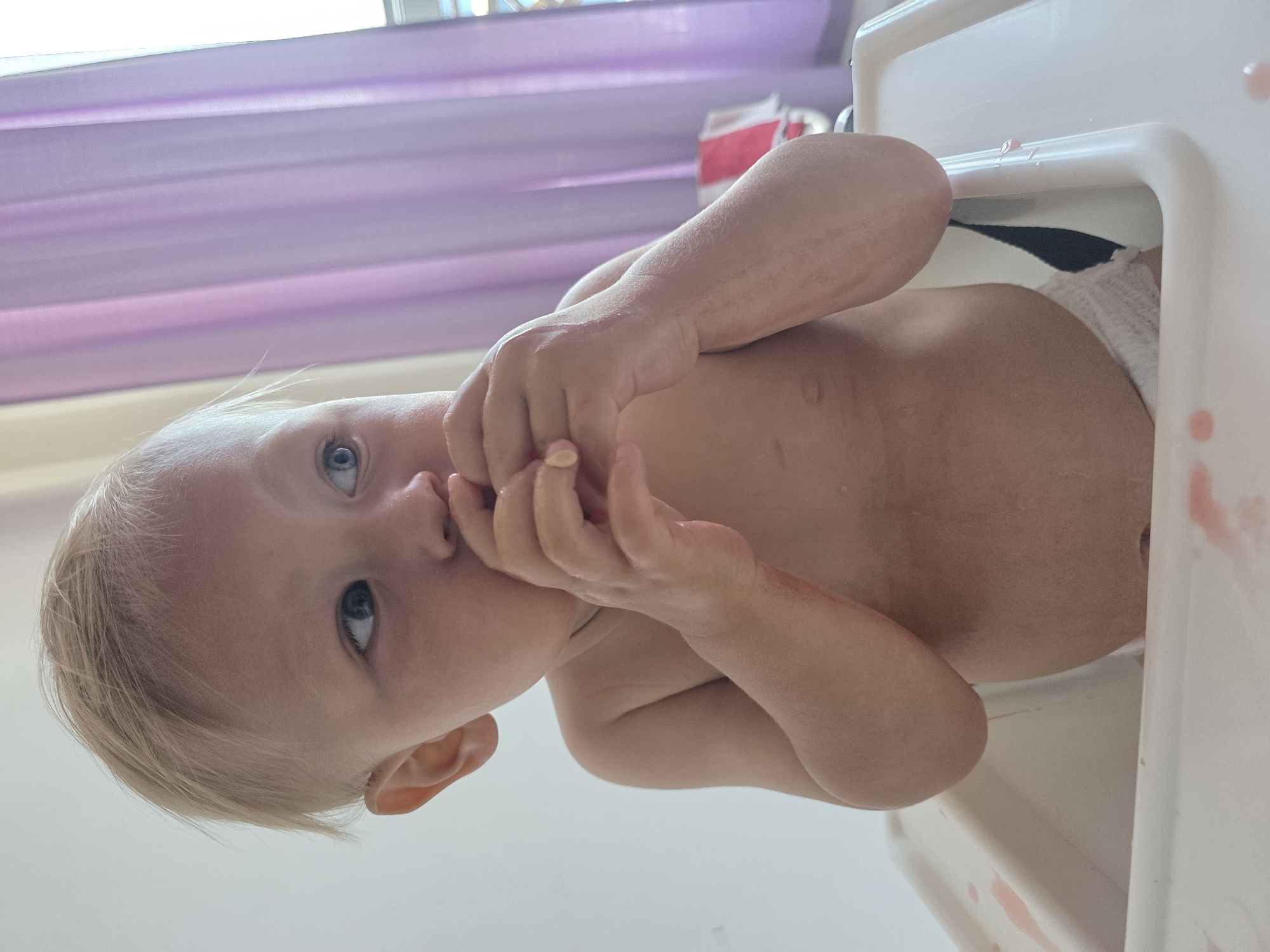 A young, fair-haired child, wearing only a white diaper, sits in a white high chair. The child has a light complexion and blue eyes, and is looking to the right with their hands near their mouth. The high chair has red smudges on its tray. In the background, sheer purple curtains hang over a window. The scene appears to be indoors.