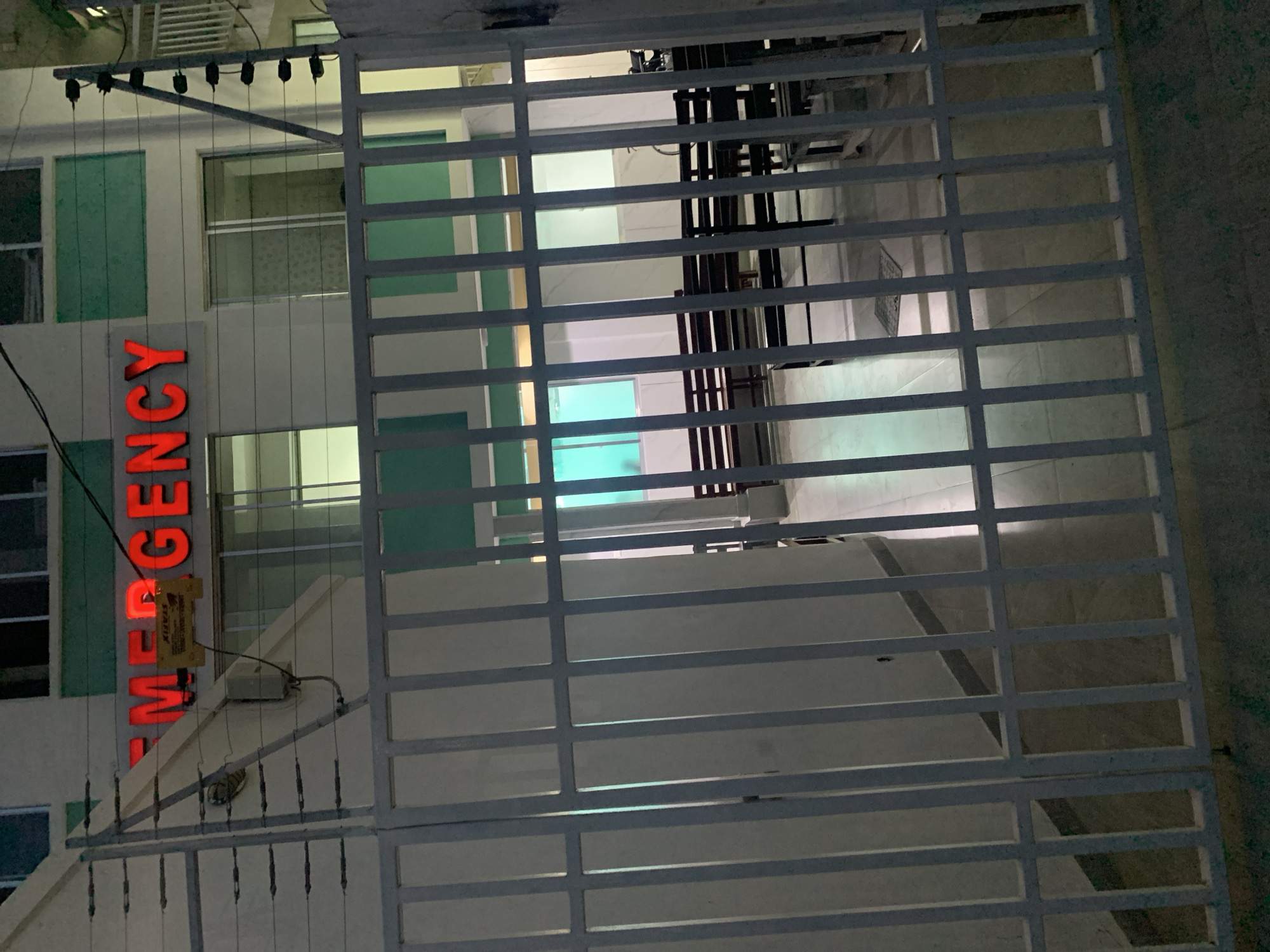 The image shows a partially obscured view of an emergency room entrance through a white metal gate. The emergency sign is illuminated in red vertical letters. The building behind the gate appears to be a multi-story structure with a white facade and green window accents. Inside, seating arrangements are visible. The location of the building is Dar es-Salaam, Tanzania.