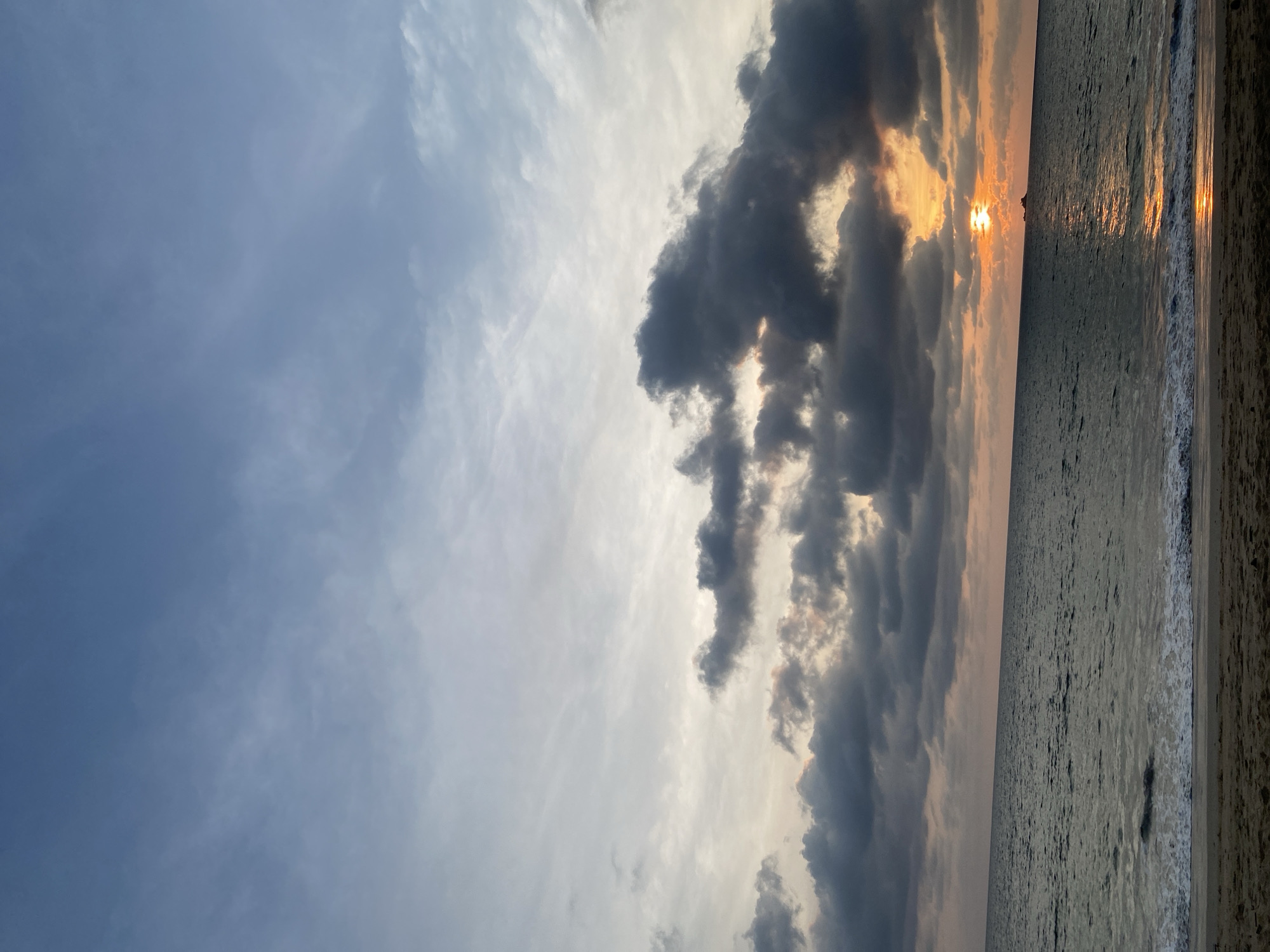 The image captures a serene sunset over the Andaman Sea in Choeng Thale, Thailand. The scene is dominated by a dramatic sky and a vast expanse of ocean.
The upper portion of the image features a dynamic sky, characterized by a blend of light and dark clouds. On the left, the sky is a soft, pale blue with delicate, wispy clouds. Towards the center and right, thick, dark grey clouds create a striking contrast, through which the vibrant, golden light of the setting sun breaks through. This illumination creates bright orange and yellow hues on the cloud edges, hinting at the sun's position low on the horizon.
Below the sky, the ocean stretches out, its surface appearing dark and rippled. A shimmering path of golden and orange light, directly reflecting the sun, extends across the water towards the foreground, indicating the sun's fiery presence. Gentle waves can be seen washing onto a narrow strip of light-colored sand along the far right edge of the frame, marking the shoreline.
The overall atmosphere is one of calm and natural beauty, typical of a tropical sunset. No human subjects, objects, or visible text are present in the image.