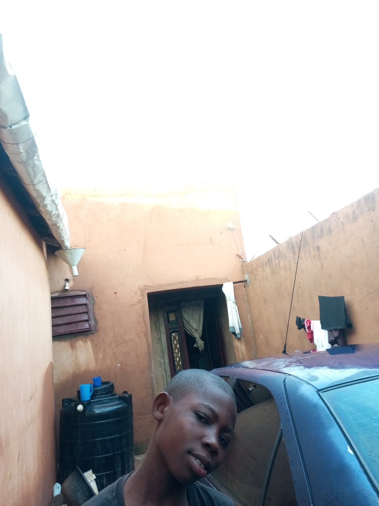 The image depicts an outdoor scene in Tempure, Nigeria, showing a young boy in the foreground. He is positioned to the right, looking towards the camera with a slight smile. Behind him, a dark-colored car is visible, with raindrops on its surface. The environment appears to be a residential area, with a large black water tank featuring two blue cups on top. Adjacent to the tank, a downspout with a funnel is attached to the wall of a building with ochre-colored plaster. A window with maroon louvers is also visible on the same wall. Further back, a doorway with decorative elements and a curtain can be seen. Clothes are hung on a line between two poles on the right side of the frame, and another set of clothes hangs near the doorway on the left. The sky is bright and overcast, suggesting a cloudy day.
