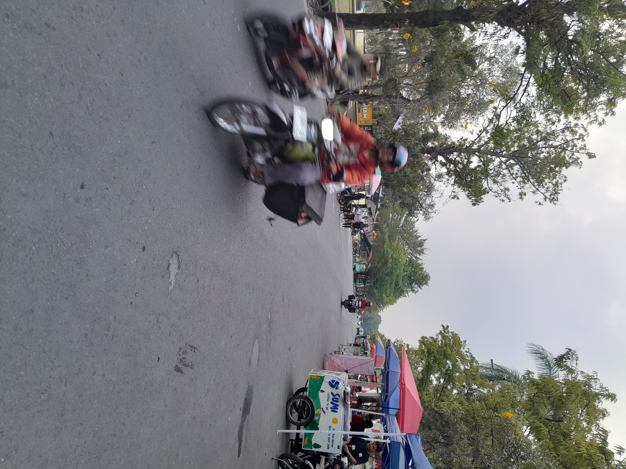 The image depicts a street scene in Slawi, Indonesia, likely captured during daytime hours. The foreground is dominated by an asphalt road with some wet patches and debris. Two motorcycles are in motion, their forms blurred due to the movement. The motorcycle on the right is ridden by an individual wearing a red jacket and a helmet, with a black storage box mounted behind them. Another motorcycle to the left is ridden by a person wearing a green jacket and a cap.
Along the right side of the road, a row of market stalls is set up, covered by colorful awnings, predominantly red and blue. One stall has a cart with "Sumi coffee to go" branding, featuring illustrations of greenery and a sun. People are visible behind the stalls, some attending to customers or preparing goods.
Trees line both sides of the street, with lush green foliage. In the background, more stalls and parked motorcycles are visible, suggesting a lively commercial area. The sky is overcast, contributing to a diffused lighting condition. The overall impression is of a busy street with mobile vendors and local commerce.