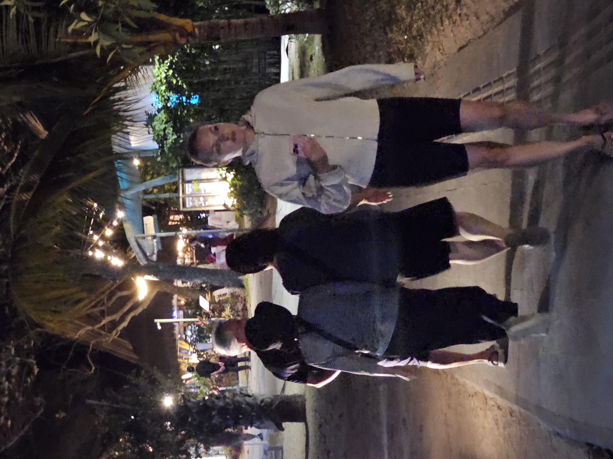 In Denpasar, Indonesia, a group of people walk along a paved pathway at night, illuminated by decorative string lights. In the foreground, a woman wearing a light-colored jacket and dark shorts is holding a pink smartphone in her right hand. To her left and slightly behind her, two individuals are walking away from the camera; one is wearing a dark top and dark shorts, and the other is wearing a light-colored top and dark pants. Further in the background, several other people are visible, as well as palm trees and buildings, suggesting a tropical or resort-like setting. The pathway appears to be bordered by sand and vegetation.