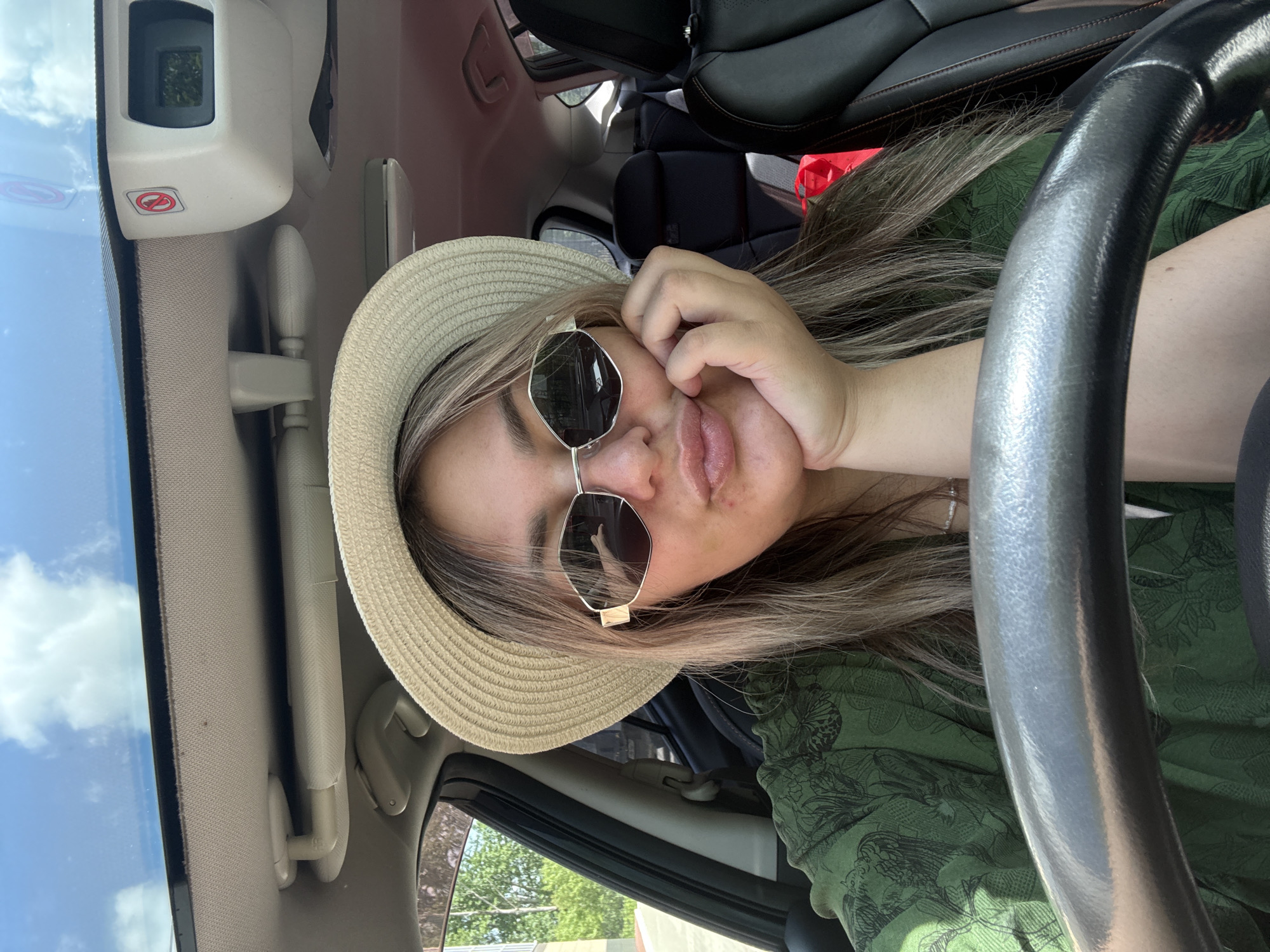 A young woman with blonde hair, wearing a straw hat and sunglasses, sits in the driver's seat of a car, leaning her chin on her hand. She is wearing a green, patterned shirt and has her lips pursed. The car interior is visible, including the steering wheel, dashboard, and seats. Outside the car, trees and a blue sky with clouds can be seen. The location is not specified as Birobidzhan, Russia.