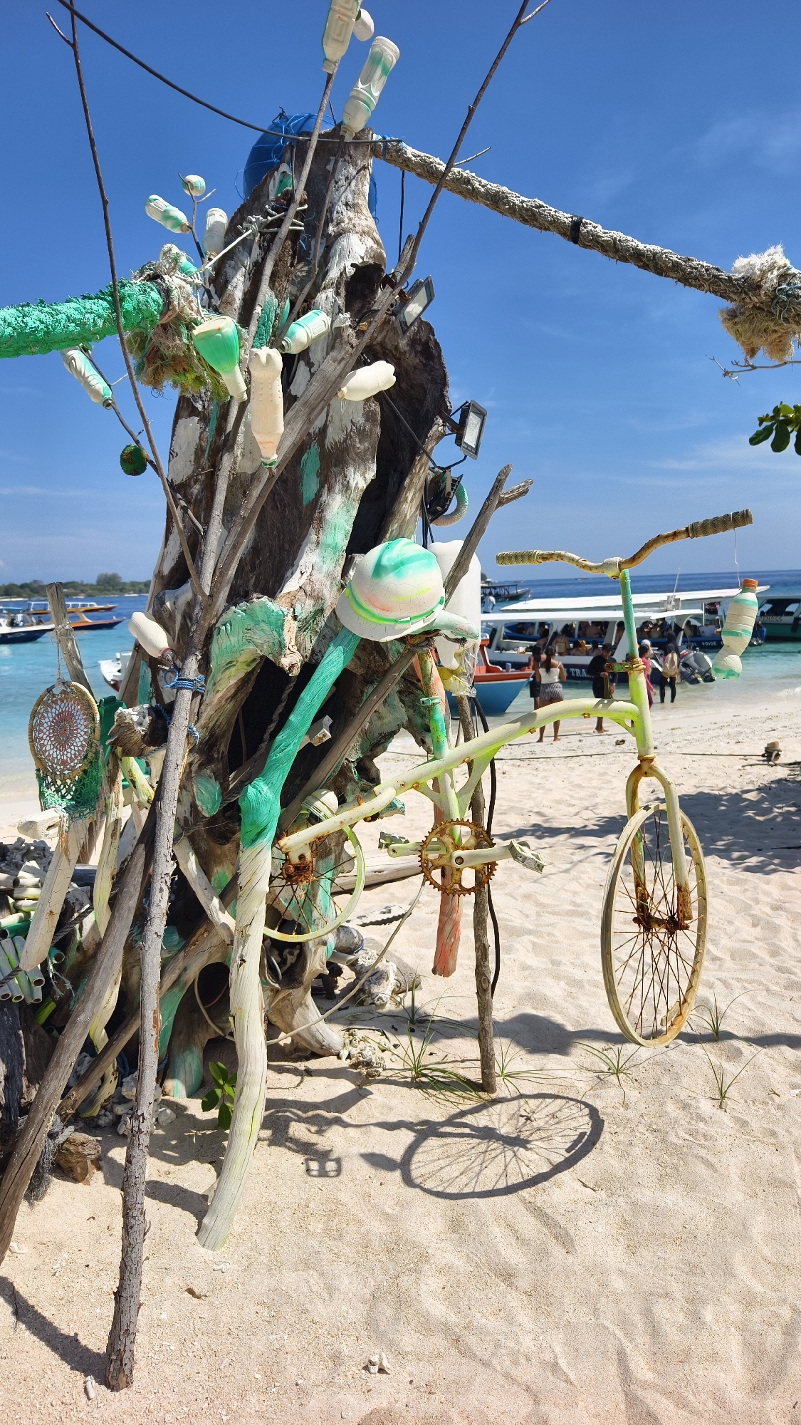 A sunny, outdoor shot at an Unknown location in Indonesia shows a sandy beach scene. The focal point is a driftwood structure adorned with various items including plastic bottles, rope, a dream catcher, and an old, green-painted bicycle. The structure appears to be a piece of art or installation. In the background, the clear turquoise water is visible, with boats and people standing on the shore. The sky is bright blue with a few wisps of clouds.