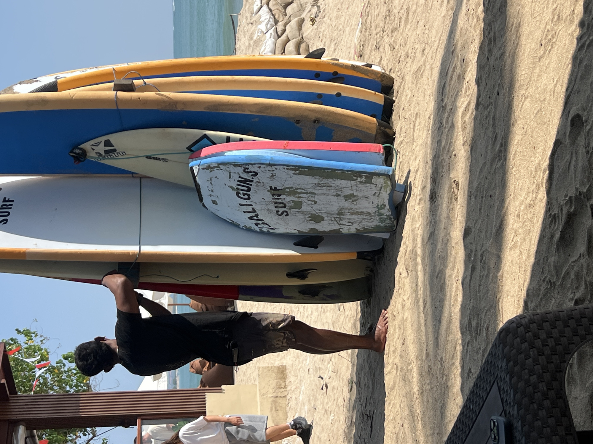 This image captures a scene on the beach in Kuta, Indonesia, a popular tourist destination known for its surf spots. In the foreground, a man, likely a surf instructor or employee, is seen from the waist down, wearing a black t-shirt and patterned shorts. He is barefoot and is in the process of tying a rope around a stack of surfboards. The surfboards are leaning against a wooden structure, possibly part of a beach cafe or rental shop, with the ocean visible in the background. The sand is a light beige, with some footprints and shadows cast by the sun. The surfboards vary in color and design, with some displaying text that reads "BALI GUN.S SURF." Other people are partially visible in the background, including one person in a white t-shirt and shorts, suggesting a lively beach environment. The overall atmosphere is casual and summery, indicative of a beach resort area.