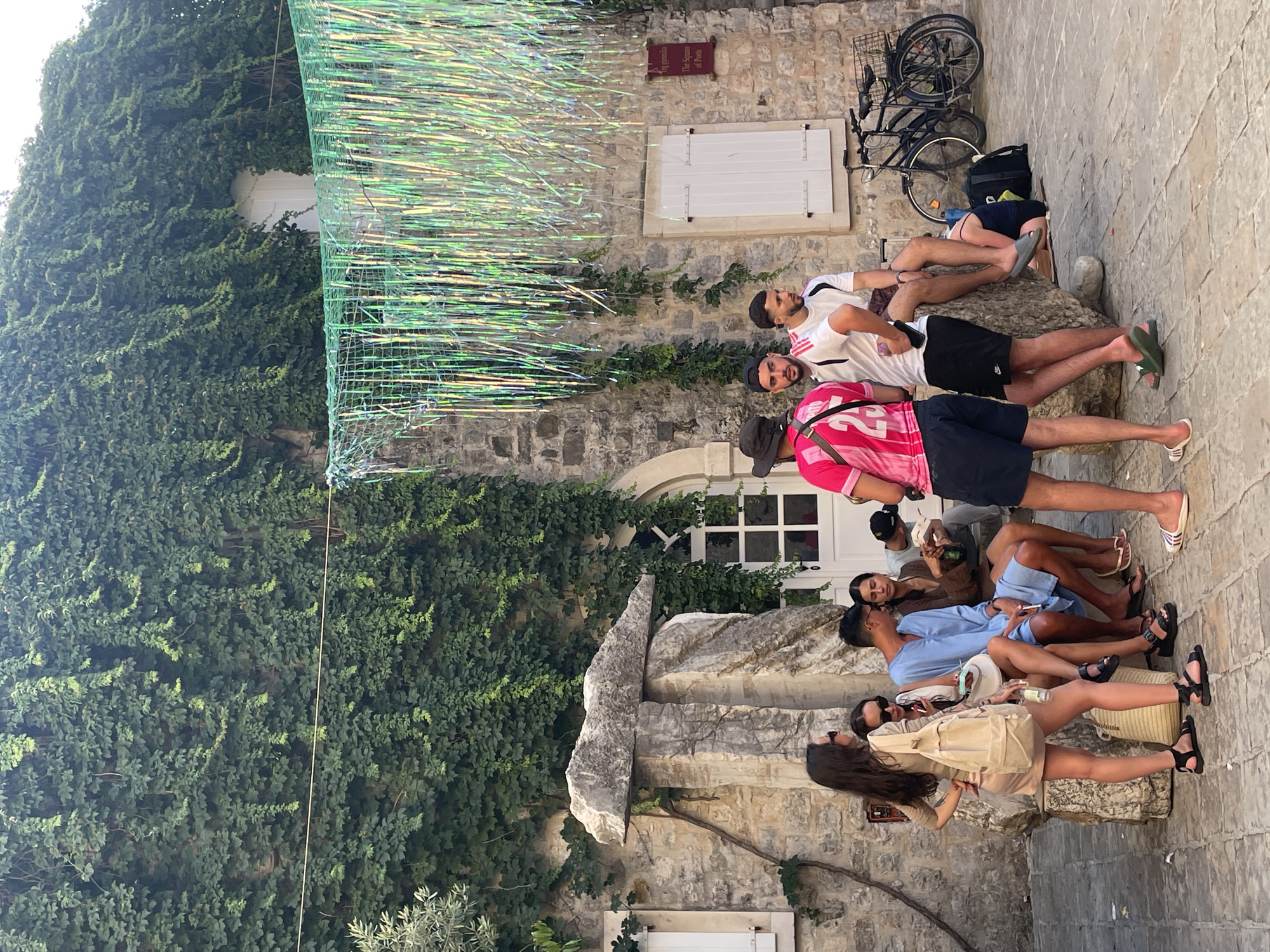 A group of young adults are gathered outdoors, appearing to be taking a break or socializing. The scene is set against a stone building with an ivy-covered wall. In the background, a bicycle is parked, suggesting an urban or historical setting. Above the people, a decorative element made of green netting and shimmering, iridescent strands hangs, possibly part of a temporary installation or decoration.
The individuals are casually dressed, some in t-shirts and shorts, others in dresses and light clothing, indicating warm weather. They are seated and reclining on what appears to be a textured stone surface or steps. Some are looking at each other, engaged in conversation, while others are looking away, perhaps at their phones or in different directions. The overall mood seems relaxed and informal.
A small sign with text is visible on the stone wall. While the text is partially obscured and not entirely clear, it seems to read "The Square of Poets." This, along with the architecture and the general ambiance, hints at a location in a historic town or a tourist area, possibly in Budva, Montenegro, as suggested by the location context. The lighting suggests it is daytime, likely late morning or afternoon, with bright sunlight casting shadows.
