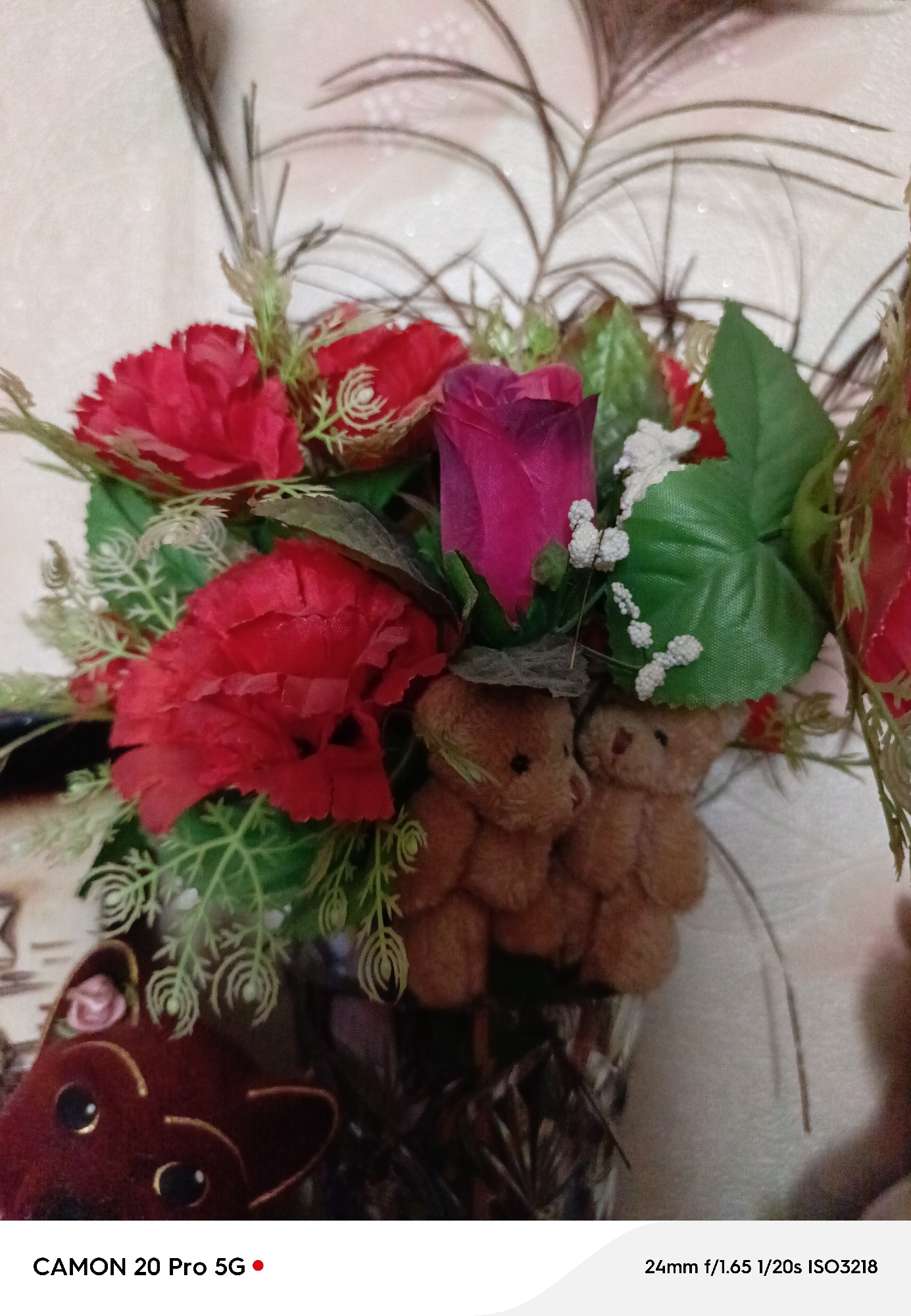 A close-up, slightly unfocused, indoor shot features a bouquet of artificial red roses and a single pink rose, interspersed with green artificial leaves and small white floral accents. Two small, brown teddy bear figures are positioned in the lower center, partially obscured by the flowers. To the left, a dark red object with large black eyes, resembling a stylized animal face, is visible. The background is a textured, light-colored wall with a subtle sparkle effect. The image displays camera information at the bottom: "CAMON 20 Pro 5G" and "24mm f/1.65 1/20s ISO3218." The scene appears to be a decorative arrangement within a home or indoor setting. There is no information in the image to determine a specific location in Yakutsk, Russia.