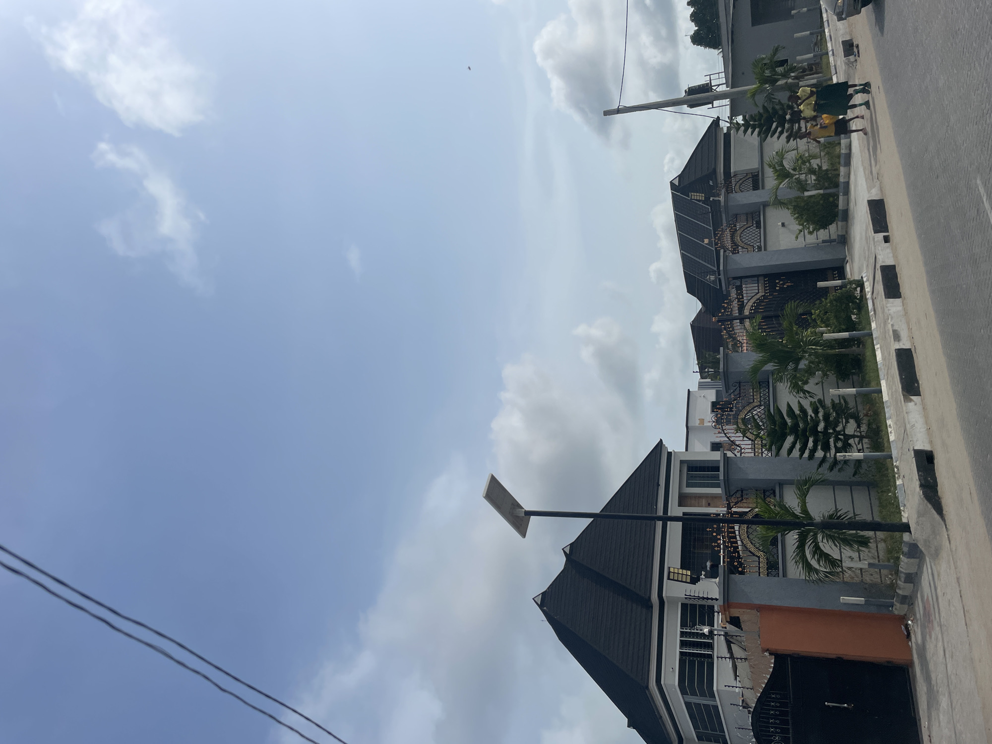 The image shows a residential street in Badagry, Nigeria. Several modern two-story houses with black roofs and gated entrances line the street. A group of three young people wearing yellow shirts and green skirts walk on the sidewalk in front of the houses. The sky is partly cloudy. A utility pole and electrical wires are visible in the background. A solar-powered street light stands near one of the houses. The scene appears to be taken during daylight hours.