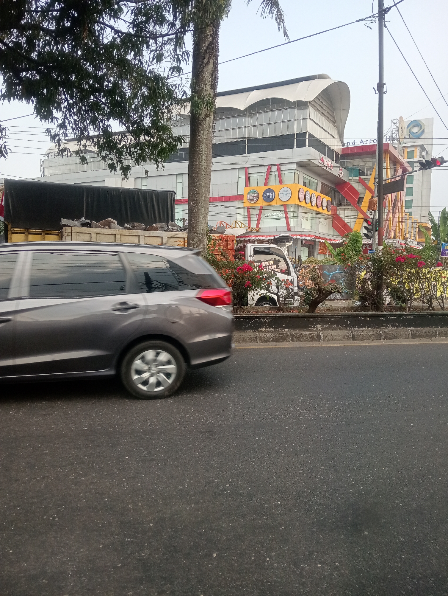 The image shows a street scene in Magelang, Indonesia, with a gray car in motion on the right side of the frame, its wheels blurred due to movement. Behind the car, a truck is visible, carrying a load of rocks. To the right of the truck, a building with a distinctive curved roof and colorful accents stands, possibly a shopping mall or commercial complex, with signage that appears to be in Indonesian. A traffic light is visible, indicating it is an intersection. Lush green trees and flowering bushes line the roadside, suggesting a well-maintained urban environment. The sky is overcast, hinting at possibly hazy or cloudy weather conditions typical of the region.