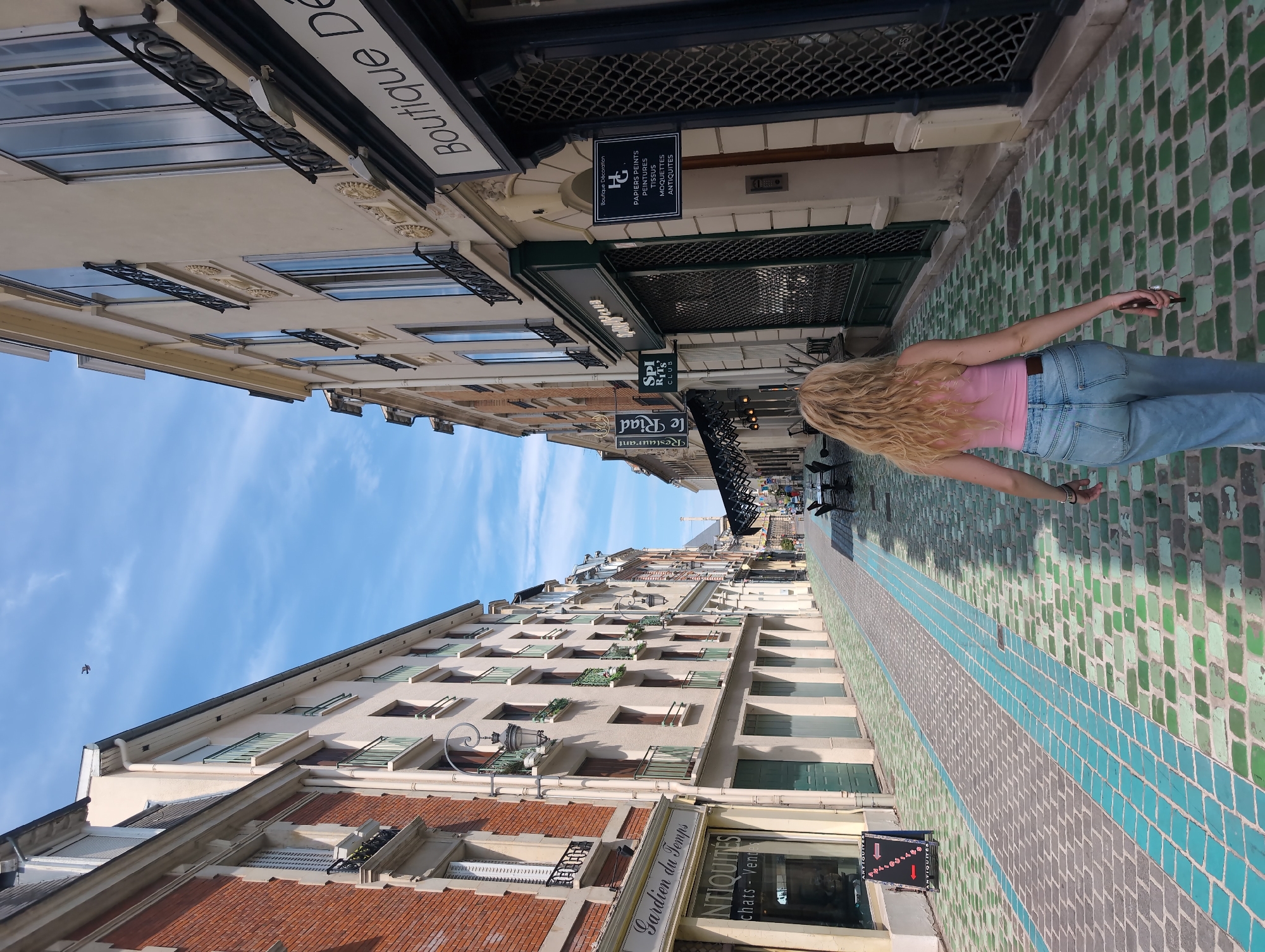 A woman with blonde, wavy hair, wearing a pink tank top and light blue jeans, walks away from the camera down a narrow cobblestone street in Reims, France. The cobblestones have a distinct pattern of grey, green, and blue tiles. On the left side of the street, a brick building has signs for "Gardien du Temps" and "ANTIQUITES". Across from it, on the right, are shops with awnings and signs, including one that reads "Boutique Décoration". The street is lined with multi-story buildings, some with wrought-iron balconies and shuttered windows. The sky above is blue with scattered clouds. In the distance, a tall tower, possibly part of a cathedral, is visible.