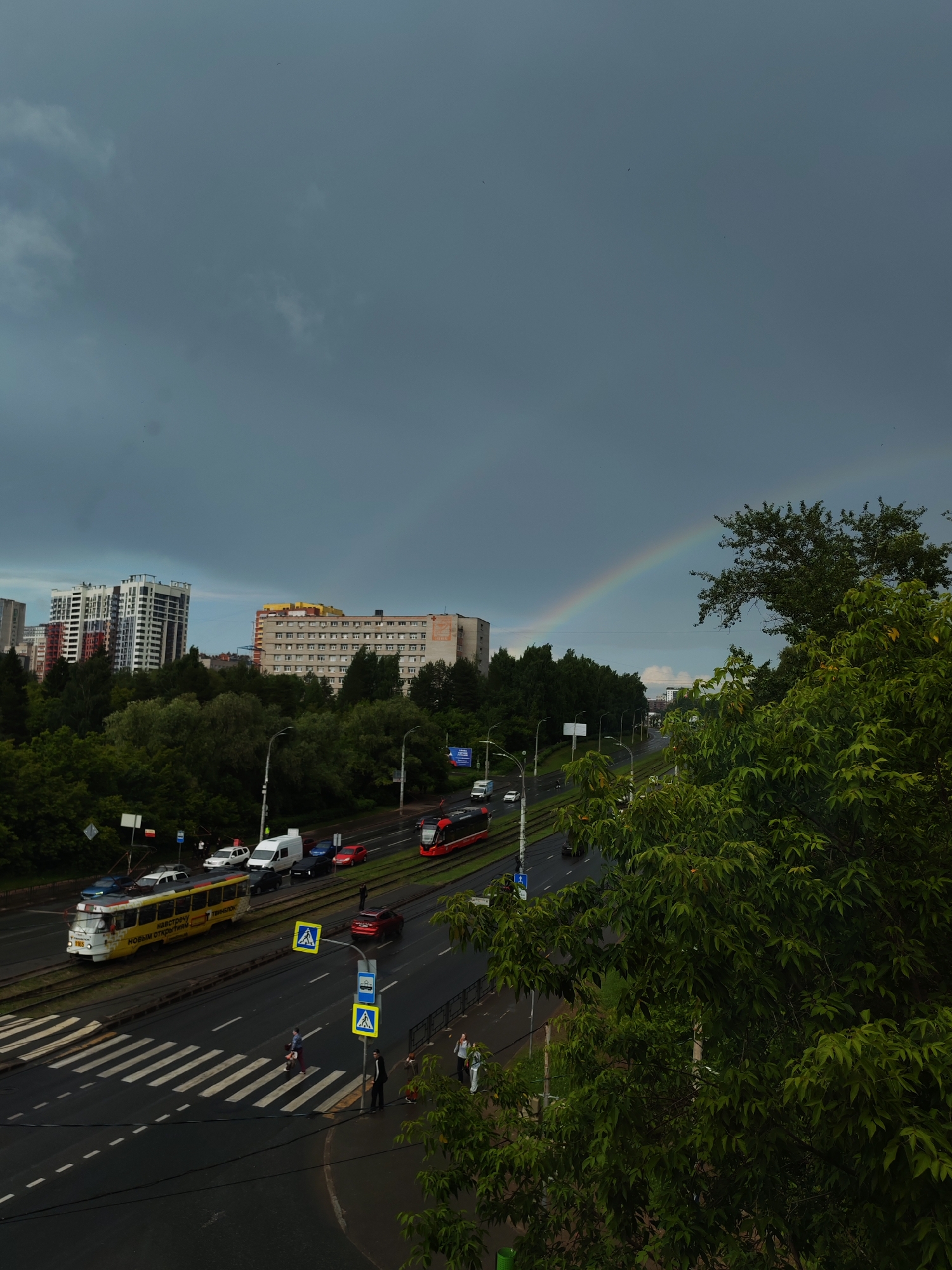 The image depicts a street scene in Izhevsk, Russia, under a cloudy sky with a rainbow faintly visible. In the foreground, lush green trees obscure the view on the right. The middle ground shows a multi-lane road with tram tracks running through it. Several vehicles are present, including a yellow tram with Russian text, a red tram, a white van, a red car, and other cars. Pedestrians are visible on the sidewalks and crossing the street at a crosswalk. Traffic lights and street signs are also present. In the background, apartment buildings of varying heights and architectural styles are visible, along with more trees. The overall impression is of a somewhat overcast day in a city with active traffic and residential areas.