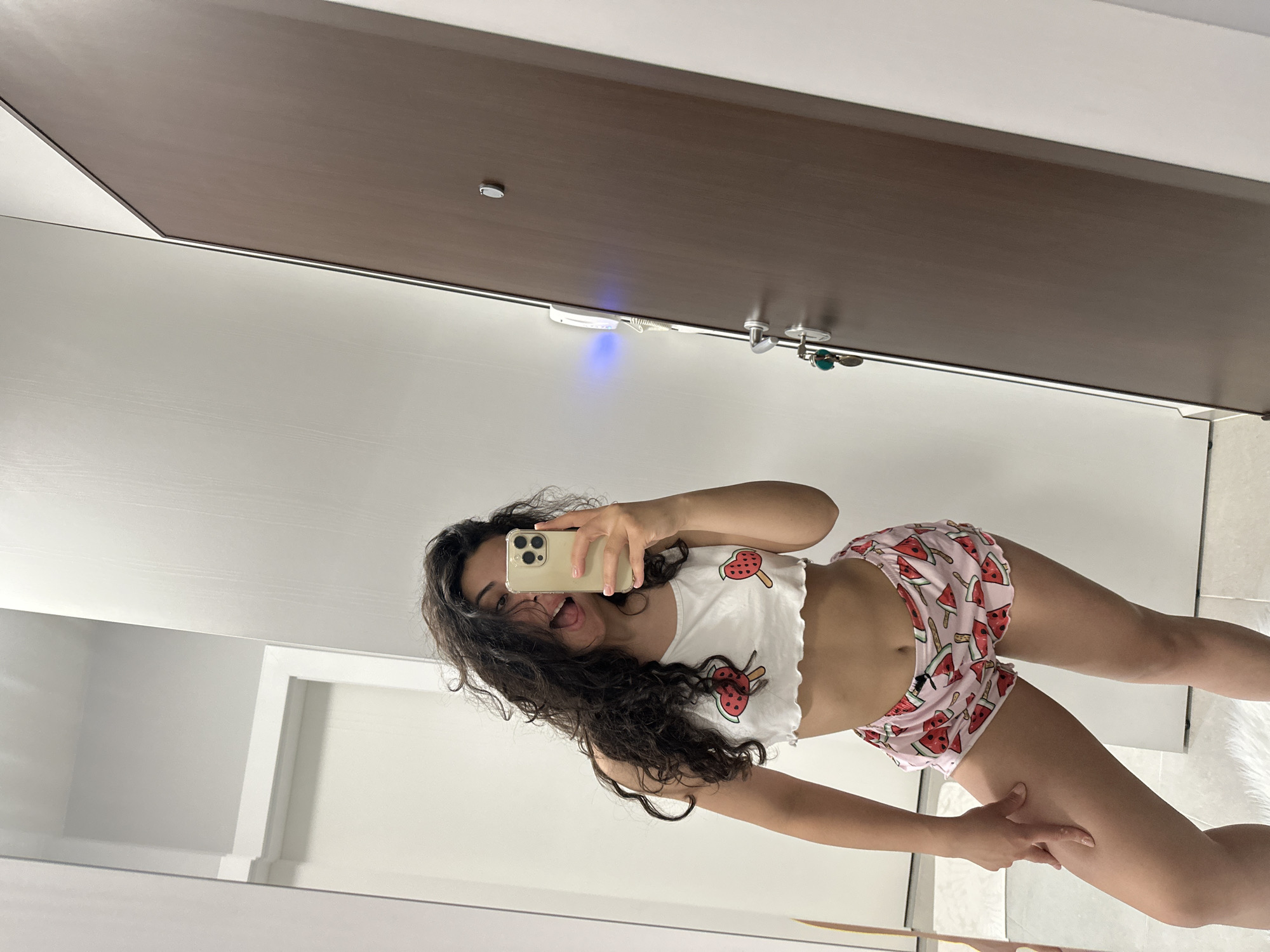 A young woman with long, dark, curly hair captures a mirror selfie indoors in the city of Unknown, Unknown. She wears a white ruffled crop top featuring two red watermelon popsicle designs and light pink shorts patterned with watermelon slices and popsicles. Her right hand holds a light-colored smartphone with a triple-lens camera system, angled towards the mirror to take the photo. Her left hand rests on her left thigh. She has a playful expression with her tongue slightly extended. The background, reflected in the mirror, shows white walls. To the viewer's left, a white doorway is partially visible. To the viewer's right, a dark brown door is equipped with a silver peephole, a silver handle, and keys hanging from it. A white intercom unit with a visible blue light emanates from its left side, mounted on the adjacent white wall. The floor beneath her appears light-colored, with a small white fluffy rug beneath her left foot.