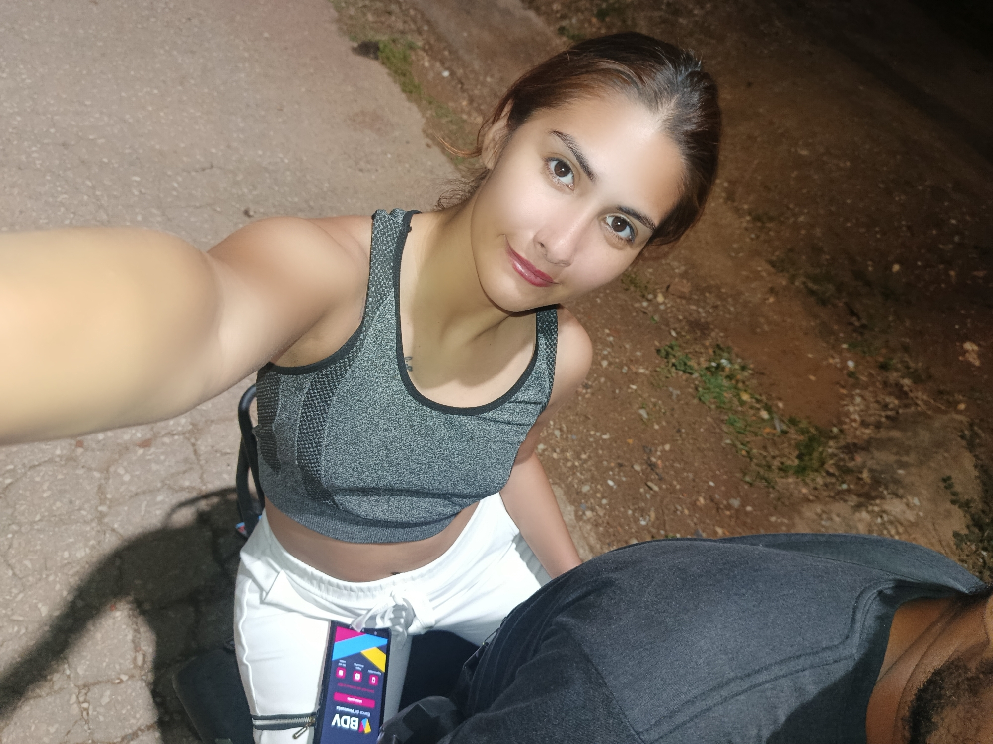 A woman is featured in a night-time selfie taken outdoors in Juan Griego, Venezuela. She is positioned in the upper center of the frame, looking directly at the camera with a neutral to slightly smiling expression. Her hair is pulled back, and she is wearing a grey athletic-style sports bra and white sweatpants with visible zippers near the ankles. Her right arm is extended, holding the camera for the selfie.
To her lower right, a partially visible person, likely male, is seated or kneeling, wearing a dark-colored top. A smartphone is resting near the woman's lap, prominently displaying an app interface. The screen shows the acronym "BDV" in large letters, with "Banco de Venezuela" written below it. Visible text labels on the screen include "Pagos", "Recargas", "Transferencias", and "Clavemóvil", indicating a banking application.
The background is dark, confirming it is night-time, with the subjects illuminated by an artificial light source. The ground beneath them appears to be a mix of paved surface and dirt, with sparse patches of grass and vegetation visible, suggesting an informal outdoor setting.
