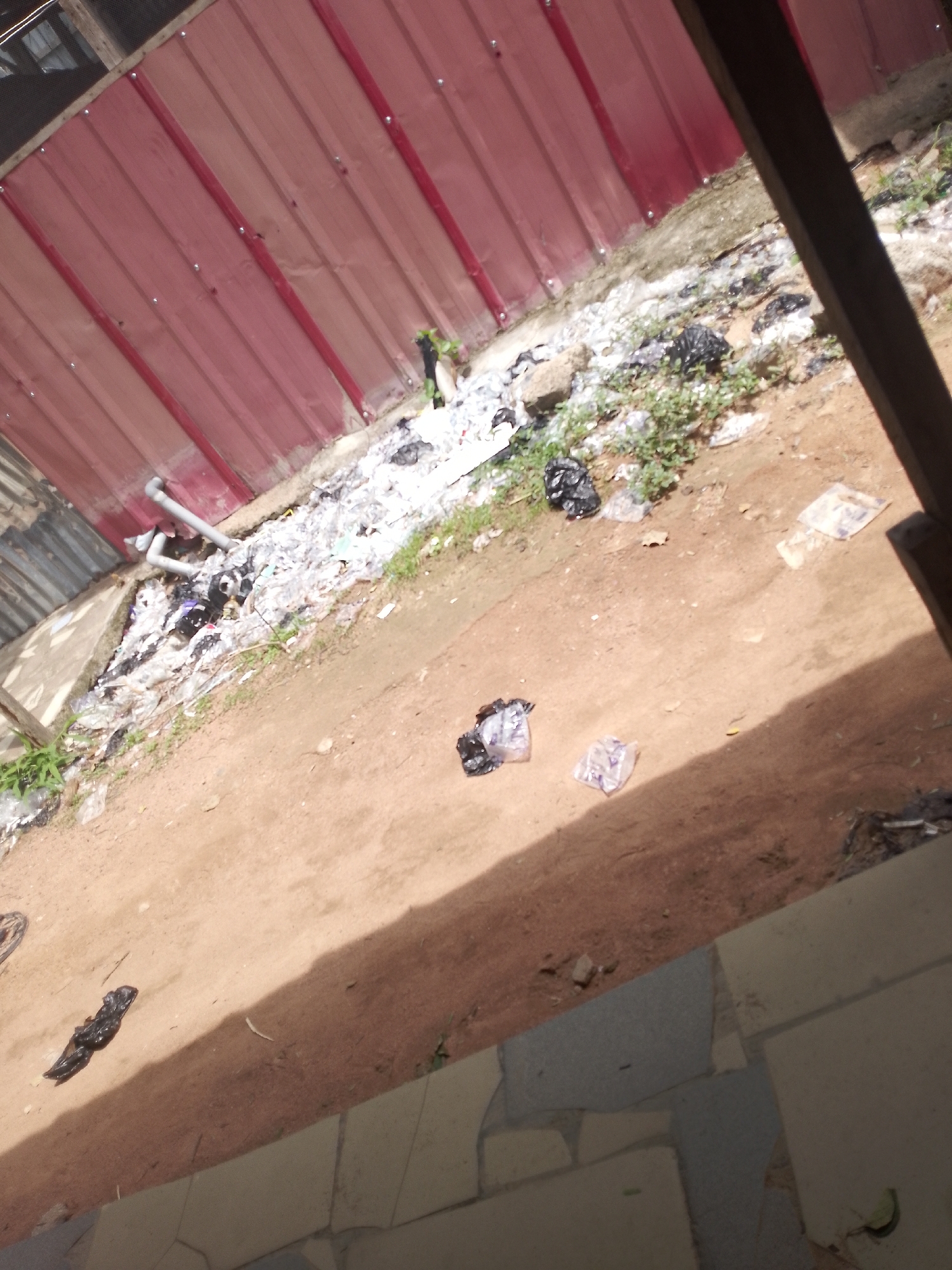 The image depicts a messy outdoor scene with several pieces of black plastic trash scattered on the dirt ground. In the background, a corrugated red metal wall runs along the top of the frame, and a patch of green vegetation is visible. The ground is a mix of dirt and some light-colored debris, possibly ash or paper. There are also a few transparent plastic bags with some contents visible, along with more black trash in the midground and foreground. A shadowed area on the right suggests a structure or an overhang, and a portion of a tiled walkway is visible at the bottom. The scene appears to be an unkempt area with a significant amount of litter.