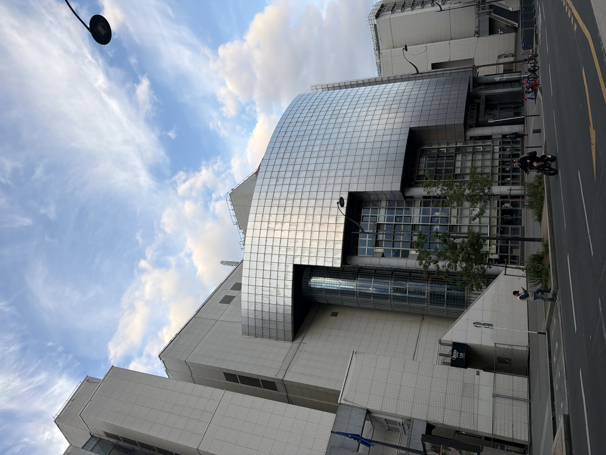 The image shows the Opéra Bastille in Paris, France, taken from street level. The building is modern, with a reflective, grid-like facade contrasting with the more textured, light-colored stone of the lower levels. The sky is partly cloudy. A few individuals are present on the sidewalk and street. One person is walking and looking at their phone. A person is riding a bicycle. Bicycles are parked on the sidewalk. A street lamp is visible.
