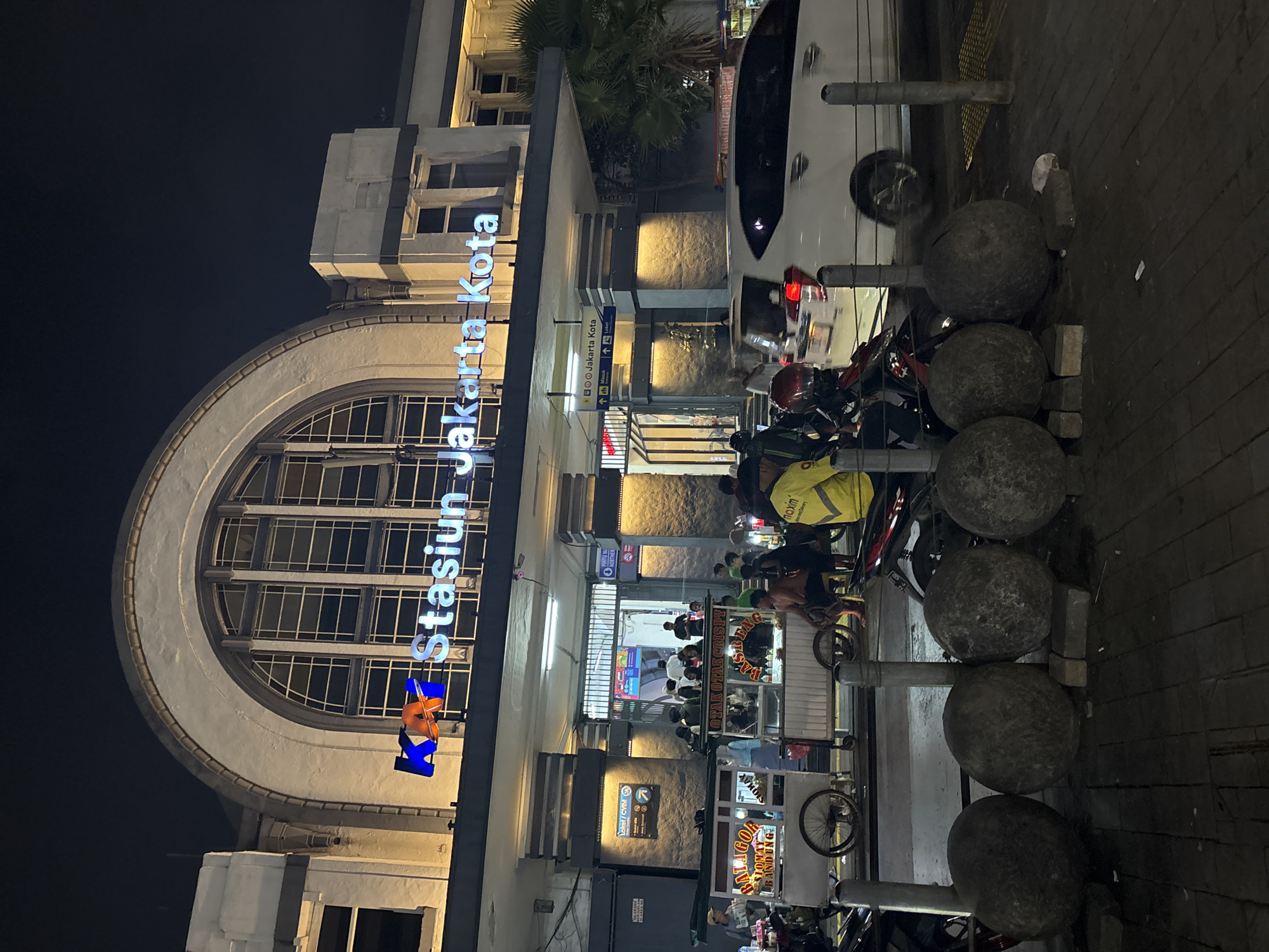 The image depicts the entrance of the Jakarta Kota train station at night, located in West Jakarta, Indonesia. A prominent sign above the entrance reads "KA Stasiun Jakarta Kota" in illuminated blue and orange lettering. The station building features a large arched window and is illuminated by warm lighting. In front of the station, several food carts are visible, with vendors and customers interacting. People are seen standing, walking, and sitting around the carts and near the entrance. Motorcycles are parked in the foreground, and a white car is driving past. The ground is paved with dark tiles, and large, rounded stone objects are placed along the sidewalk. The overall scene suggests a busy public space at night, with commercial activity and transit operations.