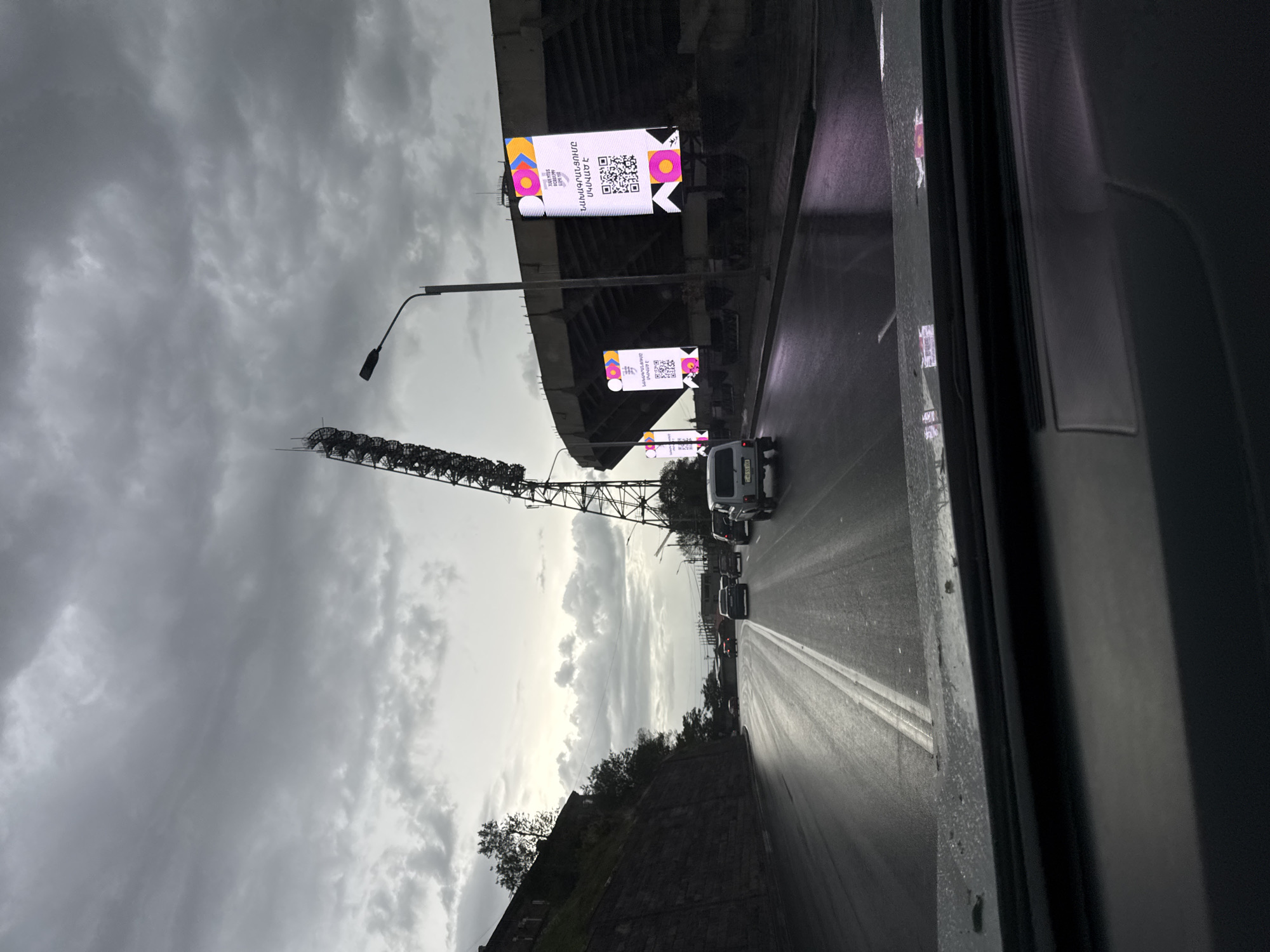 The image depicts a street scene, likely captured from inside a vehicle, in Yerevan, Armenia. The scene includes road traffic, a stadium or overpass, and advertisement billboards. The weather appears overcast with a grey, cloudy sky.
On the right side of the frame, portions of the interior of the vehicle are visible. Outside, the road is wet, with visible lane markings. Traffic includes multiple vehicles, mainly cars and vans, driving along the road.
On the left, there's an elevated, possibly stadium structure with lit advertisement billboards attached to it. The billboards feature images and QR codes. Adjacent to the structure are tall metal support structures, possibly for stadium lighting. The image also includes a tall street lamp. A stone wall is visible along the lower edge of the frame.