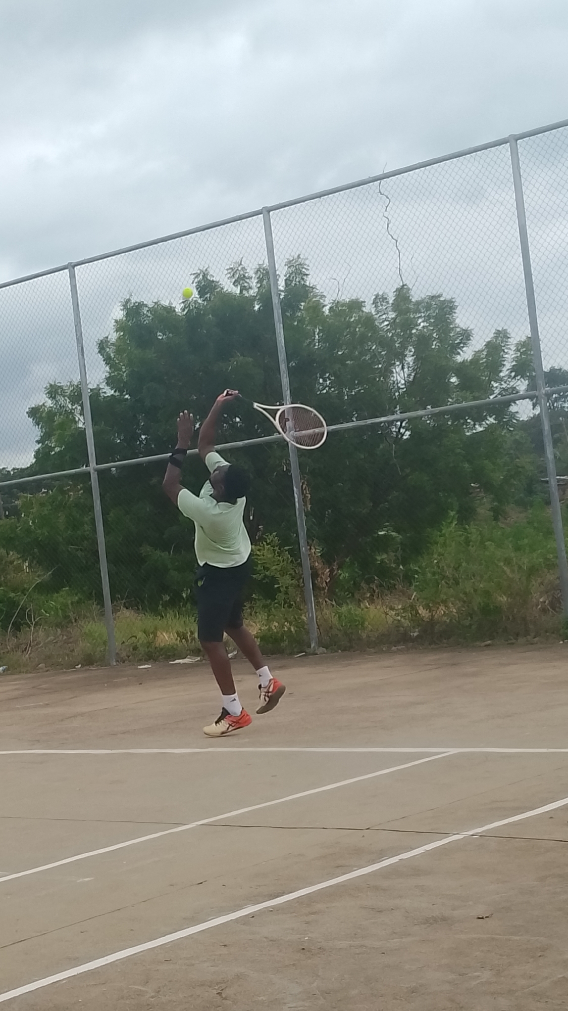 An adult male, wearing a light green short-sleeved shirt, dark shorts, white socks, and orange and beige athletic shoes, is depicted mid-air on a tennis court, performing an overhead tennis stroke. His left arm is extended upwards, and he holds a tennis racket with his right arm, positioned to strike a yellow-green tennis ball visible in the upper left quadrant of the image, near the top of the chain-link fence. The court surface is a hard, light-colored material with white boundary lines. Behind the player, a chain-link fence with metal posts stretches horizontally, separating the court from an overgrown area with numerous green trees and sparse vegetation. The sky is overcast and grey. This scene is located in Lokoja, Nigeria.