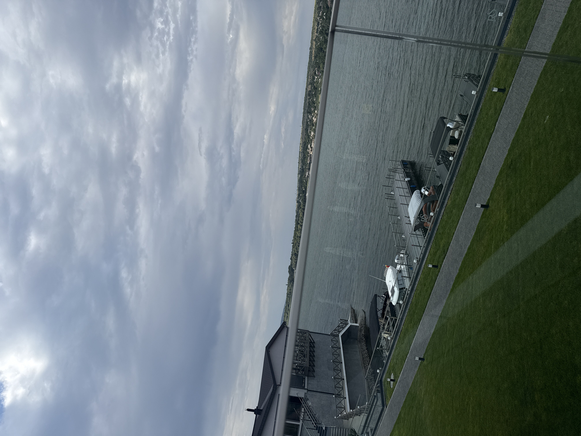 The image shows a waterside property on a cloudy day. In the foreground, there is a well-manicured green lawn with a paved walkway. A glass railing runs along the edge of the property, overlooking a body of water. A modern building with a dark roof and grey walls is situated on the left side of the image, featuring a balcony and external stairs leading down to the water. Several boats are docked at a pier extending into the water. The background reveals a distant shoreline with trees and scattered buildings, suggesting a settled area. The sky is overcast with dramatic grey clouds. The scene is serene and appears to be a resort or residential area. The location is identified as Oxentea, Moldova.