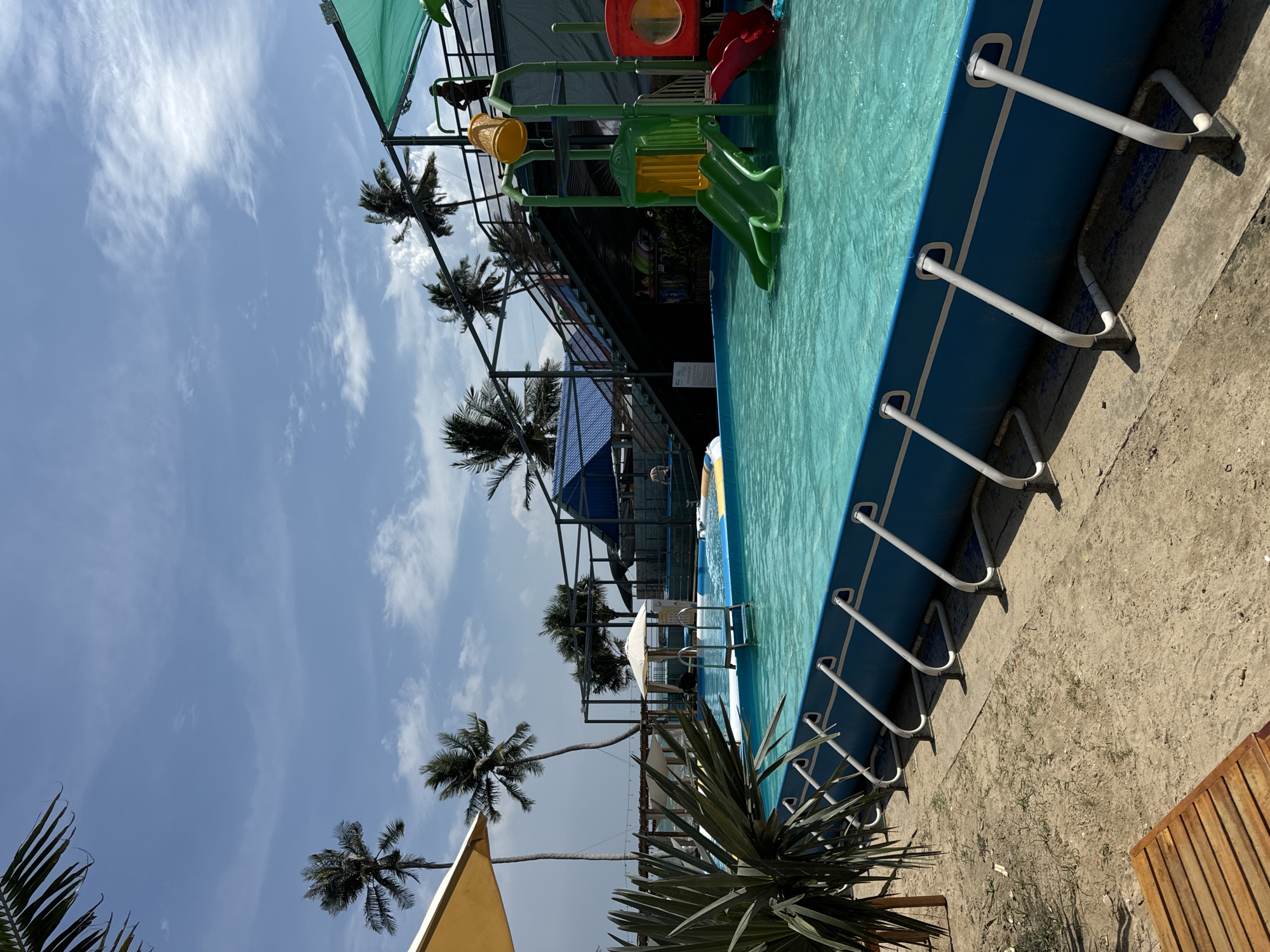 This is a medium shot of a water park on a sunny day, likely located in Mũi Né, Vietnam. The foreground is dominated by a bright blue, above-ground swimming pool filled with clear turquoise water. White metal supports hold up the sides of the pool, and a wooden lounge chair is visible in the lower left corner. To the left of the pool, a large, spiky green plant and a yellow umbrella stand out against the sandy ground and the ocean in the distance.
In the middle ground, a children's water play structure with slides in green and red is situated within the pool. A person is visible on a platform above the structure, wearing a dark outfit and a hat. Stairs lead up to this platform, and a large blue-roofed building with a darker metal frame is visible behind it.
The background features several tall palm trees swaying gently, suggesting a tropical coastal location. The sky is a vibrant blue with scattered white clouds. The overall scene conveys a sense of leisure and recreation.