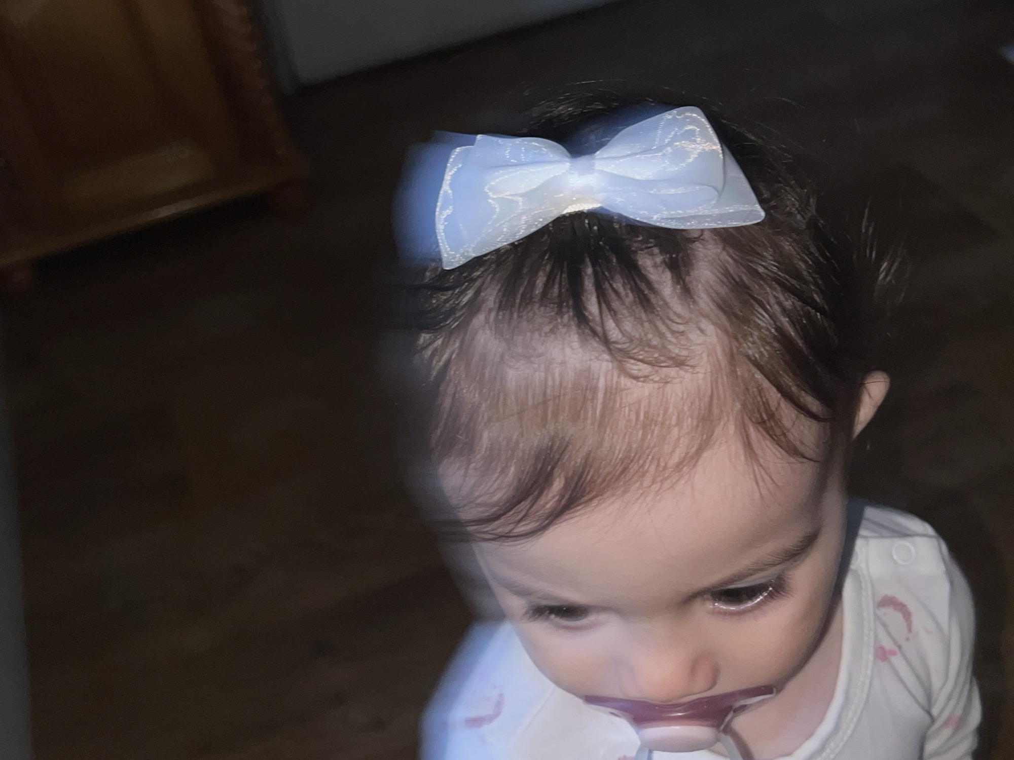The media file presents a close-up, dimly lit portrait of a baby, likely a girl, situated indoors in Elena, Bulgaria. The baby has dark, fine hair, with a prominent, sheer white or very pale blue hair bow featuring a subtle shimmering or embroidered pattern, positioned centrally on her head. Her skin tone is fair, and she has dark, expressive eyes with long lashes, gazing downwards intently. A purple pacifier is comfortably settled in her mouth, suggesting a moment of calm or comfort. She is wearing a white long-sleeved top or onesie, which has some faint, possibly pink, marks or patterns visible on the front.
The background is largely dark and out of focus, indicating a low-light indoor environment, possibly evening or night. In the upper left corner, a portion of what appears to be a wooden piece of furniture, perhaps a cabinet or chest with some detailed trim, is faintly visible. The soft focus and low light create a gentle, intimate atmosphere. No specific activity beyond the baby's focused gaze and use of a pacifier is clearly depicted. There is no visible text in the image.
