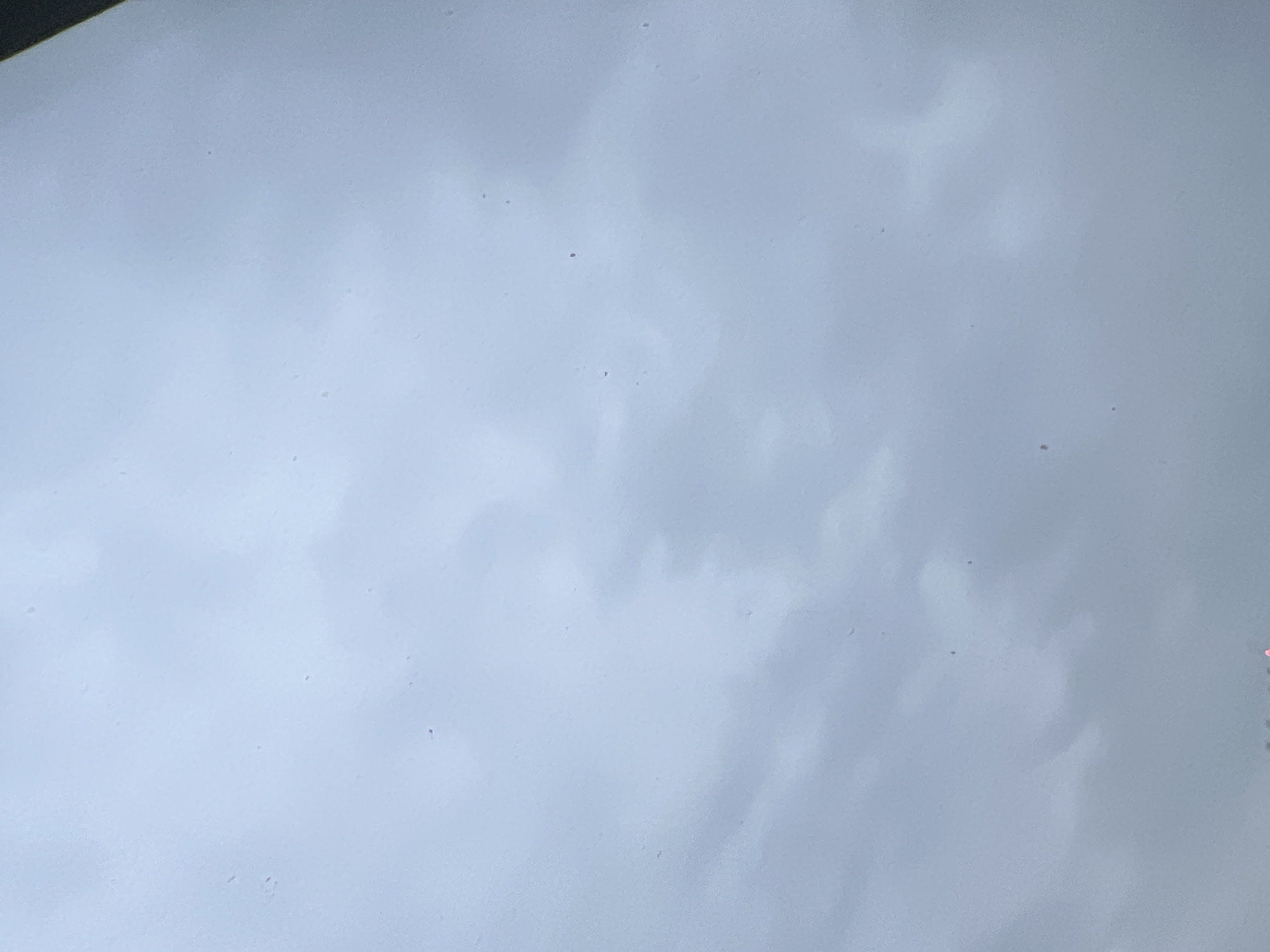 The image depicts an overcast sky with diffused light, creating a muted, grey-blue atmosphere. There are faint, wispy cloud formations visible, suggesting a cloudy day. Small, dark specks are scattered across the sky, potentially dust or minor imperfections on a lens or surface through which the photo was taken. At the very bottom edge of the image, faint, blurred lights, possibly traffic signals or building lights, are visible, suggesting an urban setting. Without additional context or identifying features, it is not possible to definitively determine the location within Warsaw, Poland.