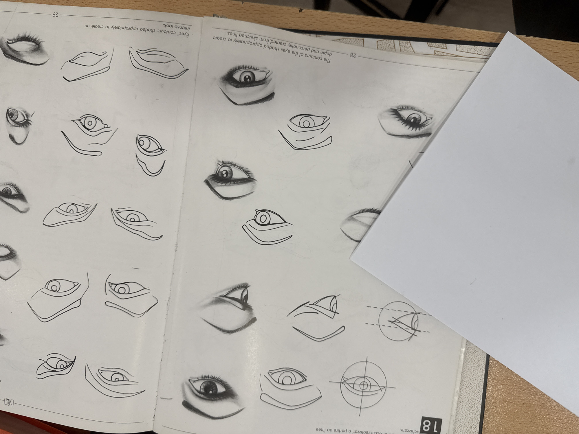 The image shows two open pages of a book on eye drawing techniques. Page 28 features eye contours shaded to create depth and personality using sketched lines, while page 29 focuses on eye contours shaded appropriately to achieve an intense look. Various eye sketches are displayed on both pages, ranging from basic outlines to more detailed and shaded representations. A blank white piece of paper partially covers the right side of page 28. The book rests on a textured surface with a dark border. The number "18" and some partially visible text in a foreign language appear in the bottom right corner. The location of the image is not discernible.