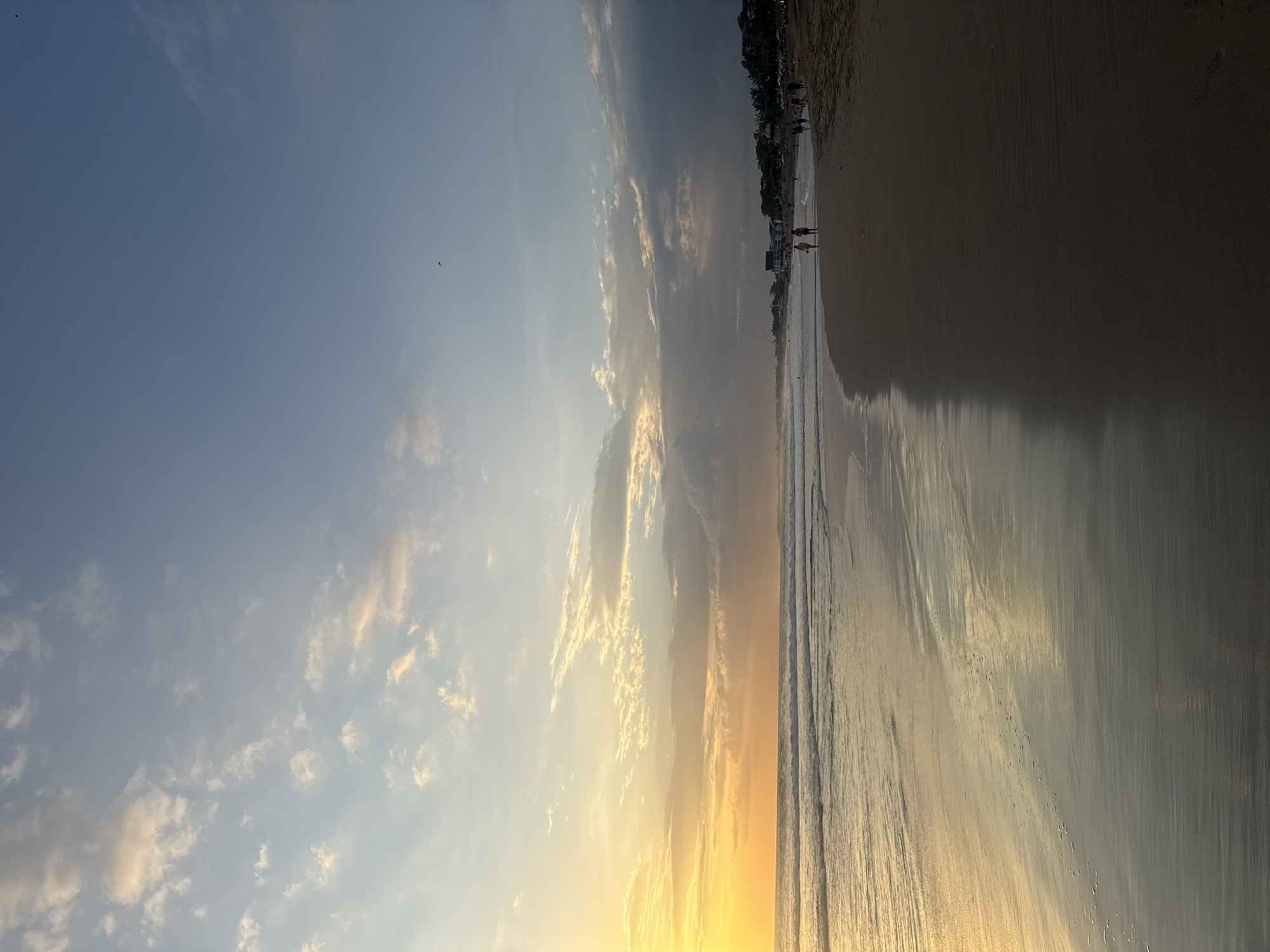 This is a beach scene in North Kuta, Indonesia, at either sunrise or sunset. The sky is divided between a clear blue on the left and golden-hued clouds on the right. The sun is positioned low on the horizon, casting a golden glow. The beach stretches across the right side of the image, with wet sand reflecting the light. The water appears calm, and there are a few people walking along the shoreline. Along the very top edge of the frame, there are trees and buildings.