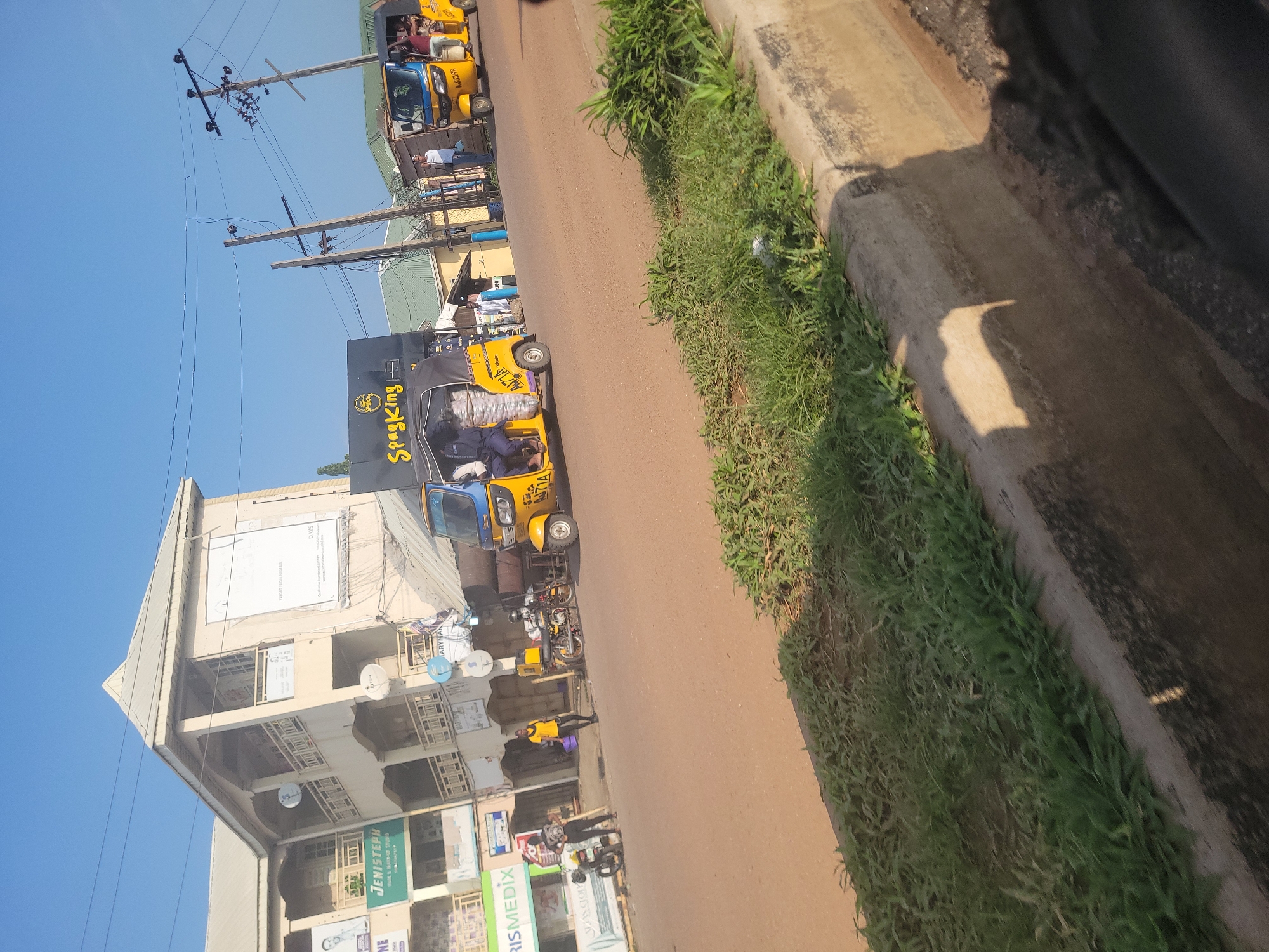 The image depicts a street scene in Lokoja, Nigeria, on a bright, sunny day. The perspective is from inside a vehicle, looking out at a bustling street. Several yellow and blue auto-rickshaws, commonly known as "keke napep" in Nigeria, are visible. Some are carrying passengers and goods, suggesting ongoing transportation activities.
One prominent auto-rickshaw has "Spagking" written on its side in bold yellow letters. Other auto-rickshaws have license plates that read "AJ71A" and "AJ3348". In the background, a multi-story building with a corrugated metal roof is visible. Signs on the building include "JENISTEPH MADE & MAKE-UP STUDIO" and "RISMEDIX".
There are also people on the street, some walking, some standing near the auto-rickshaws. Overhead, electrical wires crisscross the scene, supported by wooden poles. The overall impression is one of everyday life and commerce in a Nigerian city. The lighting suggests it is daytime, likely morning or afternoon, with clear skies.