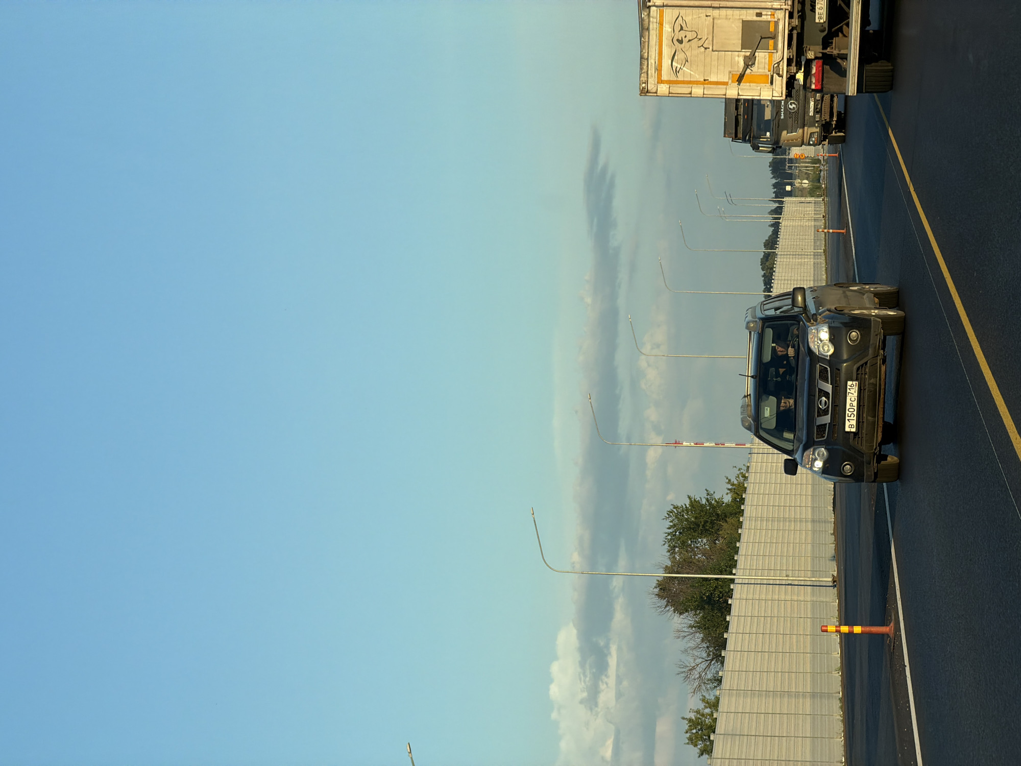 An eye-level, full shot captures a highway scene in Kaipy, Russia, under a clear, pale blue sky. The road is asphalt, marked with yellow and white lines. In the foreground, a black SUV drives in the right lane. The SUV has its headlights on, and its license plate reads "B 150 PC 716". Inside the SUV, two people are visible. To the right of the SUV, a large semi-truck with a white trailer is in the adjacent lane. The trailer has an orange and white graphic on its side. Further down the road, another semi-truck is visible. On the left side of the highway, a sound barrier wall runs parallel to the road, punctuated by orange traffic cones. Several streetlights are also visible along the highway. Trees and foliage are present behind the sound barrier.
