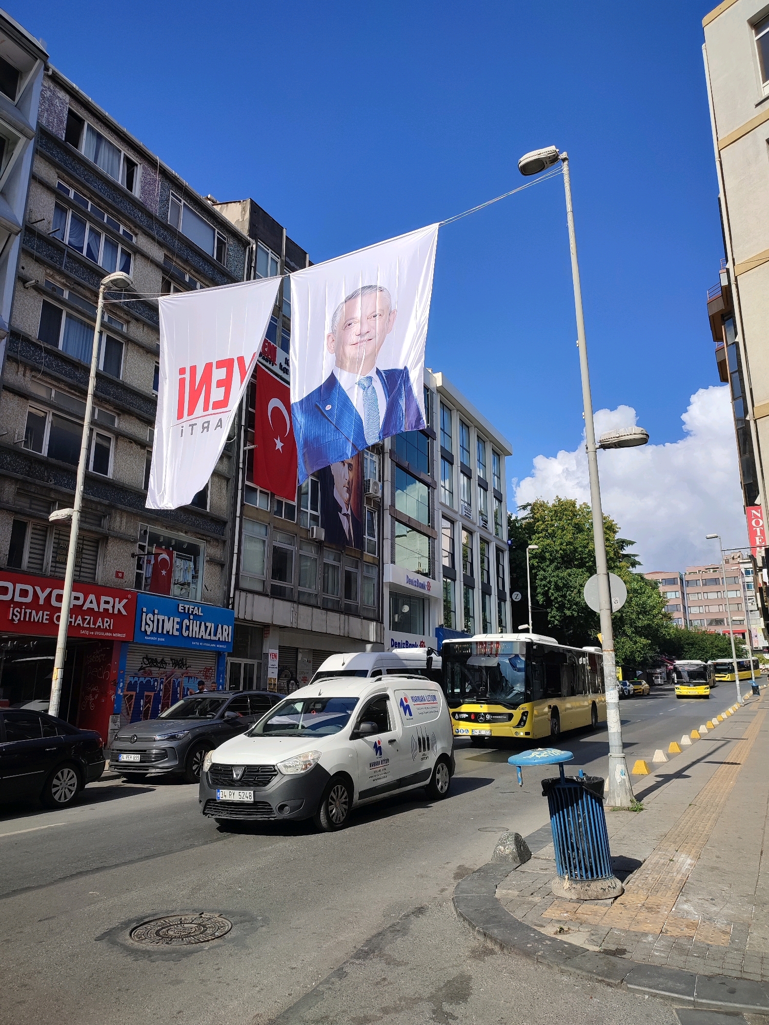 The image captures a street scene in Kadıköy, Turkey, on a bright, sunny day with a clear blue sky and scattered clouds. Large banners are prominently displayed, including one with a portrait of a man in a blue suit and tie, and another with red text that appears to say "YENI" (new). A Turkish flag is also visible hanging from a building.
The street is paved and shows signs of traffic, with several vehicles present. A white van with graphics and text is parked on the right side of the road. Behind it, a gray SUV is also parked. Further down the street, yellow buses are operating on a bus lane. Pedestrians are not clearly visible.
Buildings line the street, with various architectural styles and signage. Some buildings have multiple floors with windows. The environment includes streetlights, a trash can, and a manhole cover on the road. The overall impression is a bustling urban environment.