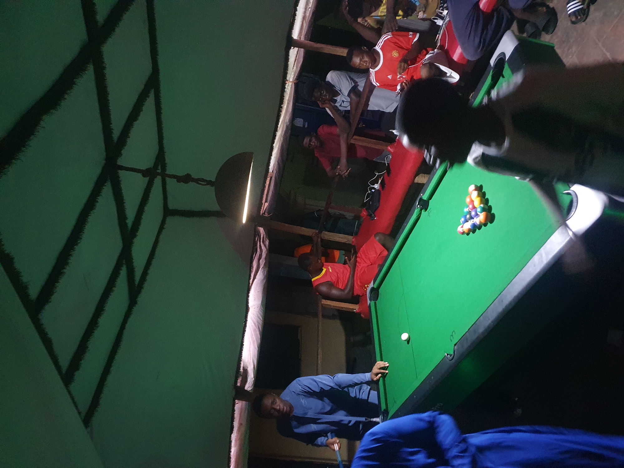 The image captures an indoor night scene in Lokoja, Nigeria, where several young men are gathered around a pool table. The setting appears to be a casual, dimly lit recreational space, possibly constructed with wooden beams and planks, giving it a somewhat informal and rustic feel.
In the foreground, a dark-skinned man wearing a blue long-sleeved shirt is actively engaged in playing pool. He is leaning over the green-felted pool table, holding a cue stick, and appears focused on his shot. A white cue ball is visible near the edge of the table, while a full rack of fifteen billiard balls, comprising solids and stripes, is neatly arranged in a triangle at the opposite end of the table, ready for play.
Several other men are observing the game. To the right of the pool table, part of another person, possibly wearing a blue garment, is visible. In the background and above the pool table, a group of men is situated on what appears to be an elevated platform or mezzanine, some leaning on a railing. Among them, one man wears a red and white striped jersey, another a light grey t-shirt, and a third a solid red shirt. They are casually watching the game unfold.
A bell-shaped light fixture hangs from the ceiling, casting a warm, bright glow over a portion of the pool table and the surrounding area, indicating it is nighttime or late evening. The ceiling above features a distinctive criss-crossing dark pattern against a green surface. No discernible text is visible on clothing, equipment, or in the environment. The overall atmosphere suggests a relaxed social gathering focused on the game of pool.