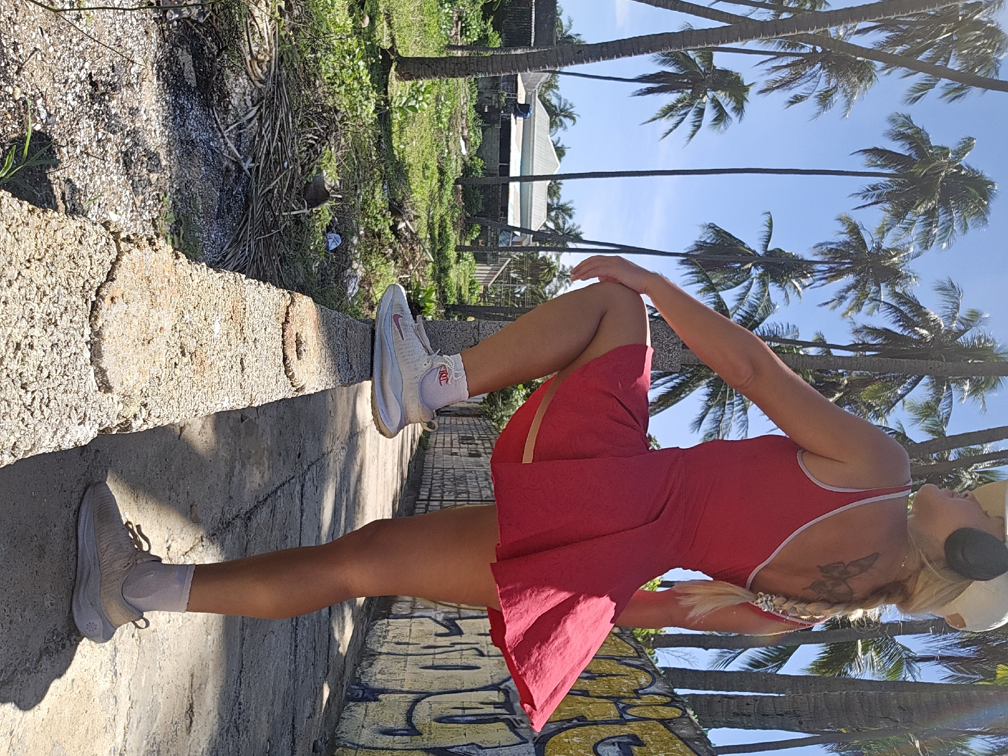 A woman stands with her right leg raised on a low concrete wall in Phu Thuy Ward, Vietnam. She is wearing a red dress, white socks, and athletic shoes. The background features palm trees, a graffiti-covered wall, and vegetation. The woman has blonde hair in a braid and is wearing headphones and a baseball cap. She has a butterfly tattoo on her upper back. Sunlight casts shadows on the ground.